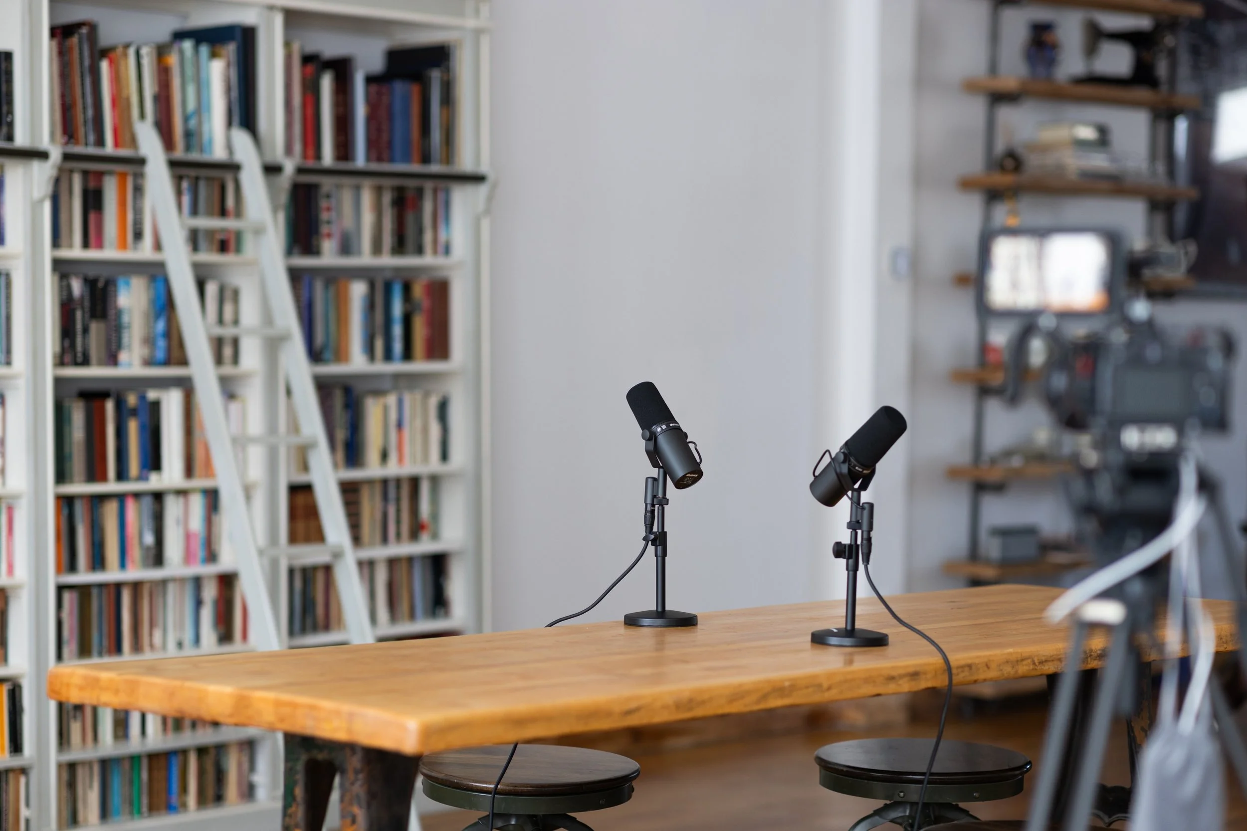 Two black microphones on a wooden table in a room with a large bookshelf filled with books, and a camera recording the setup.