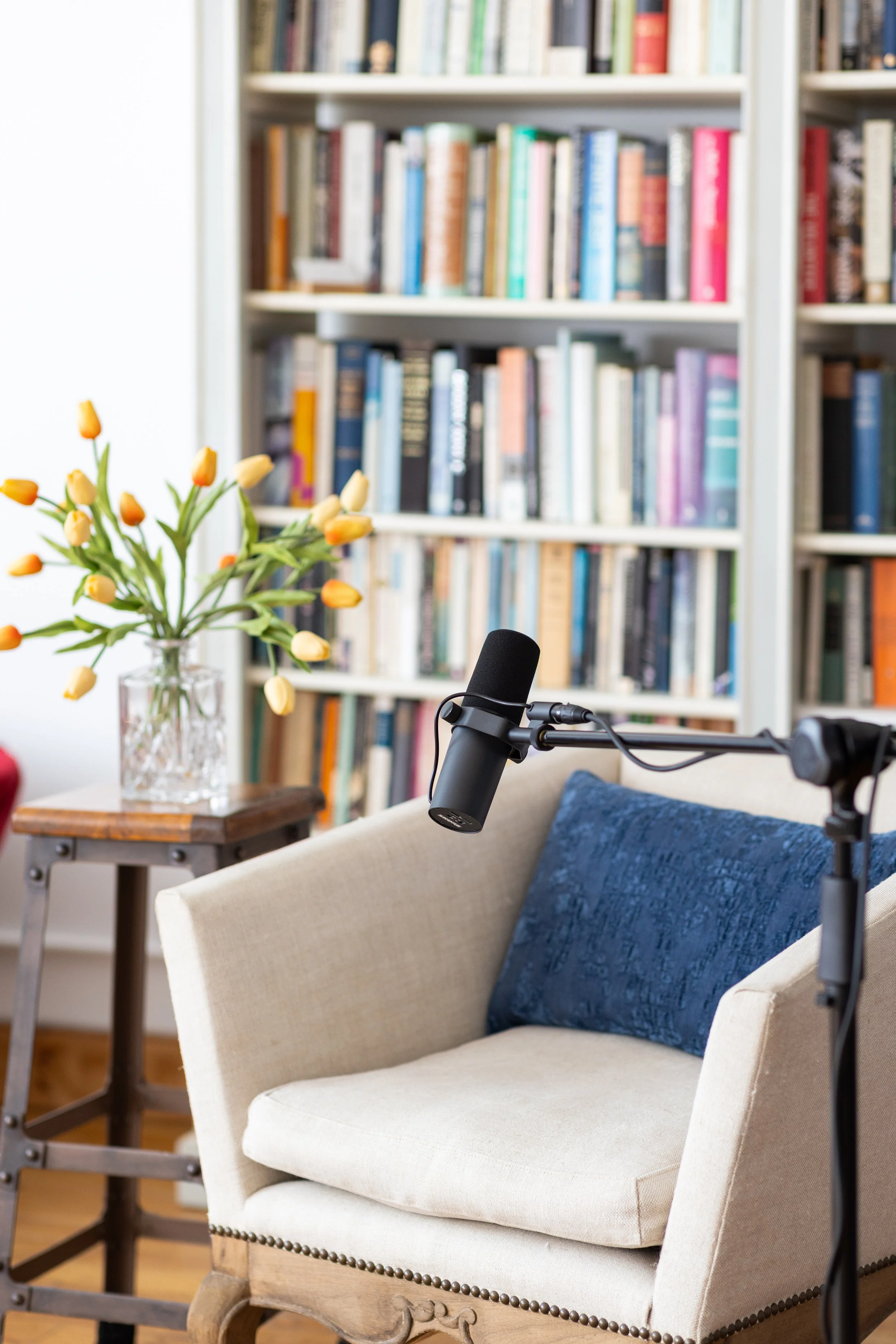 A cozy corner with a beige armchair, dark blue cushion, microphone, a side table with a vase of yellow tulips, and a bookshelf filled with colorful books in the background.