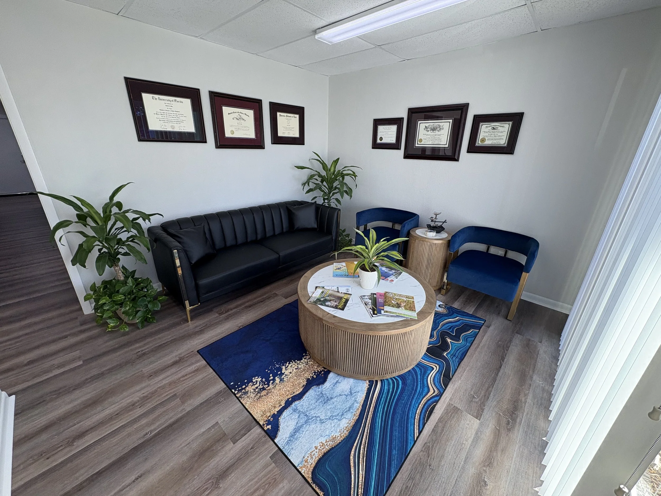 This is an image of a clean office space lobby area with a dark blue rug with gold and white accents, a black couch with gold accents, a couple plants, and diplomas within wooden frames on the light grey wall. The floor is a light brown wood.