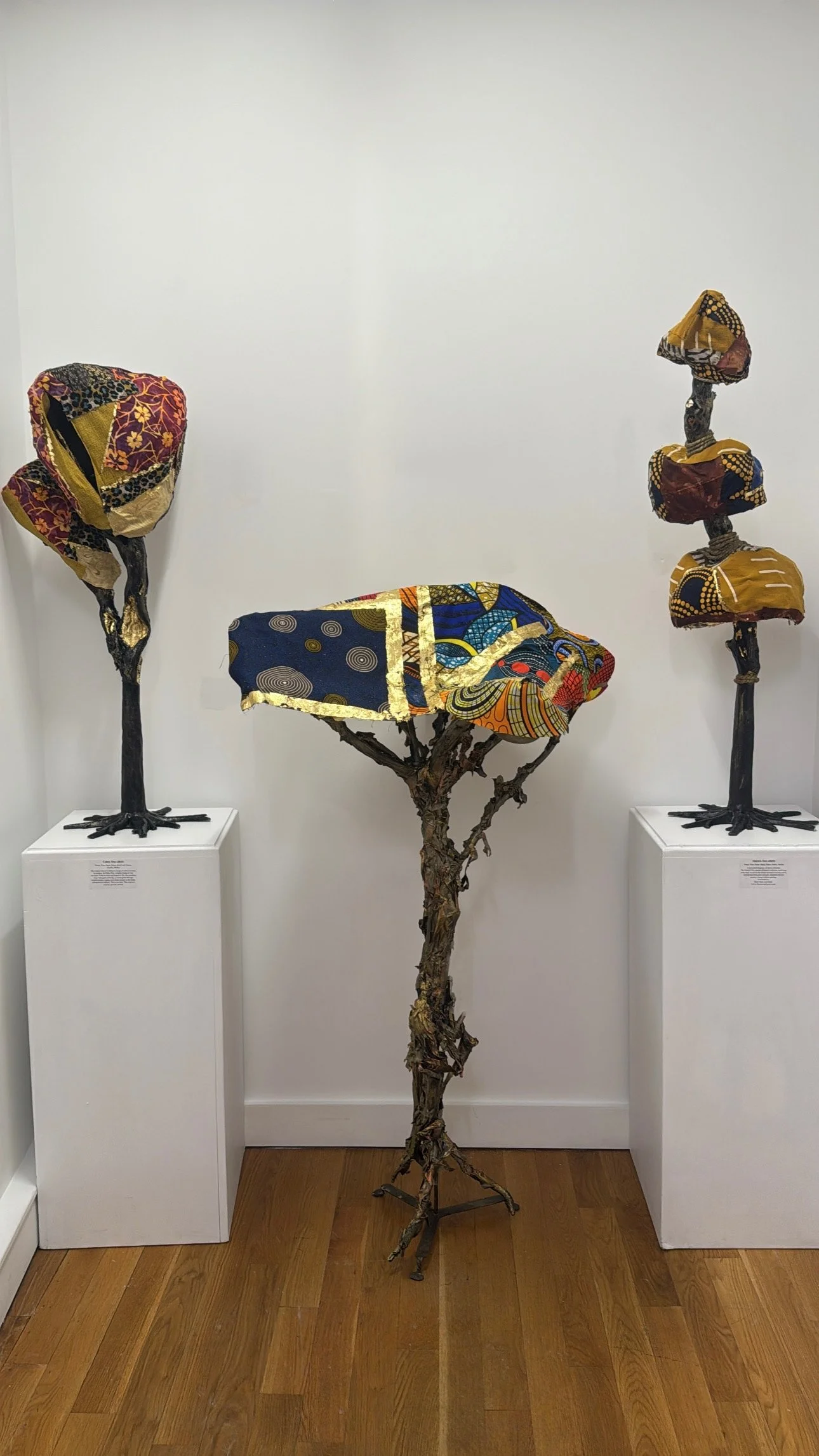 Mixed media sculpture installation featuring three trees with branches wrapped in colorful patterned fabrics, set on white pedestals against a white wall with wooden floor.