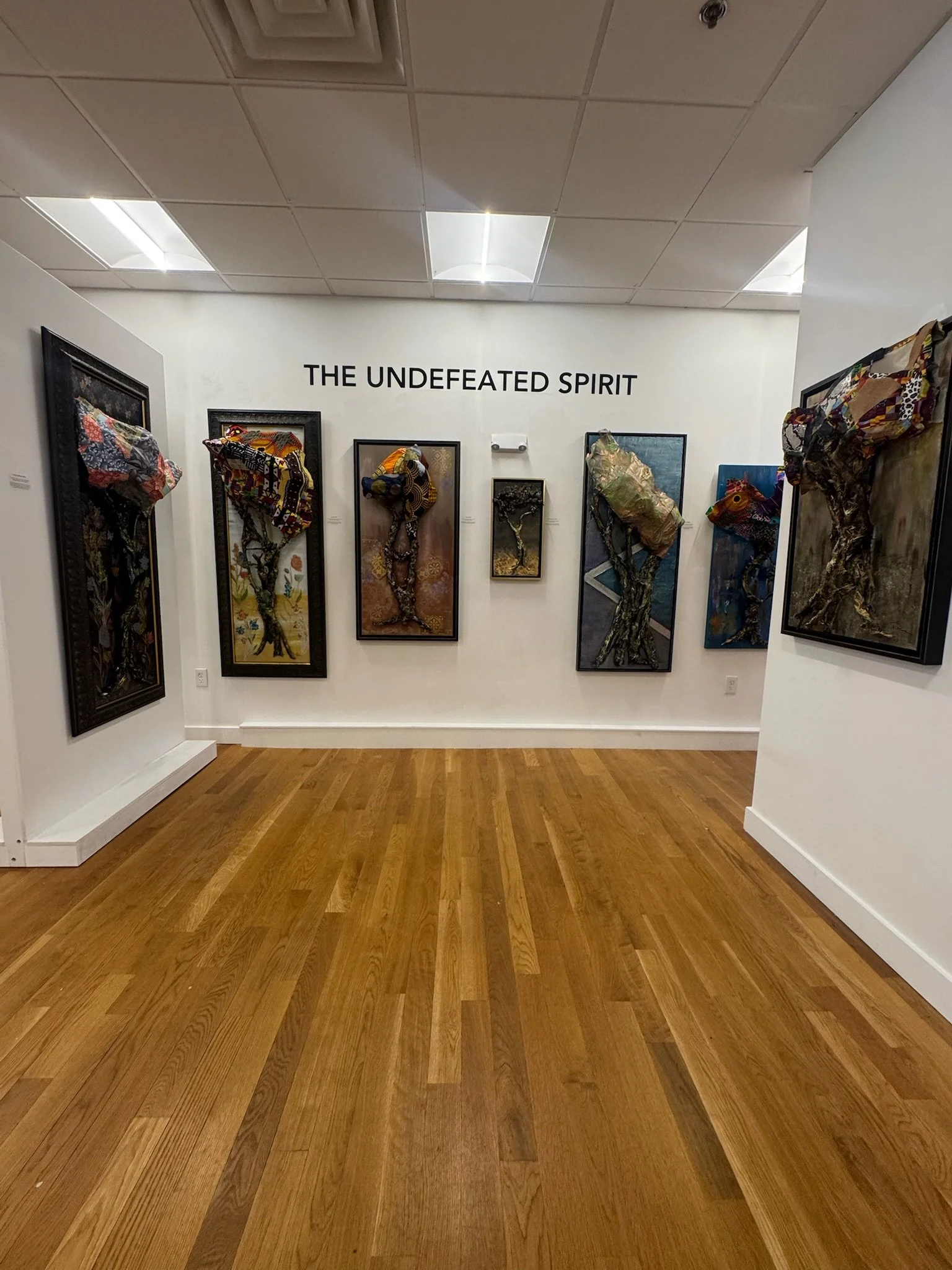 An art gallery display titled 'The Undefeated Spirit,' featuring several framed mixed media paintings of trees with fabric and textured elements on white walls, with a hardwood floor.