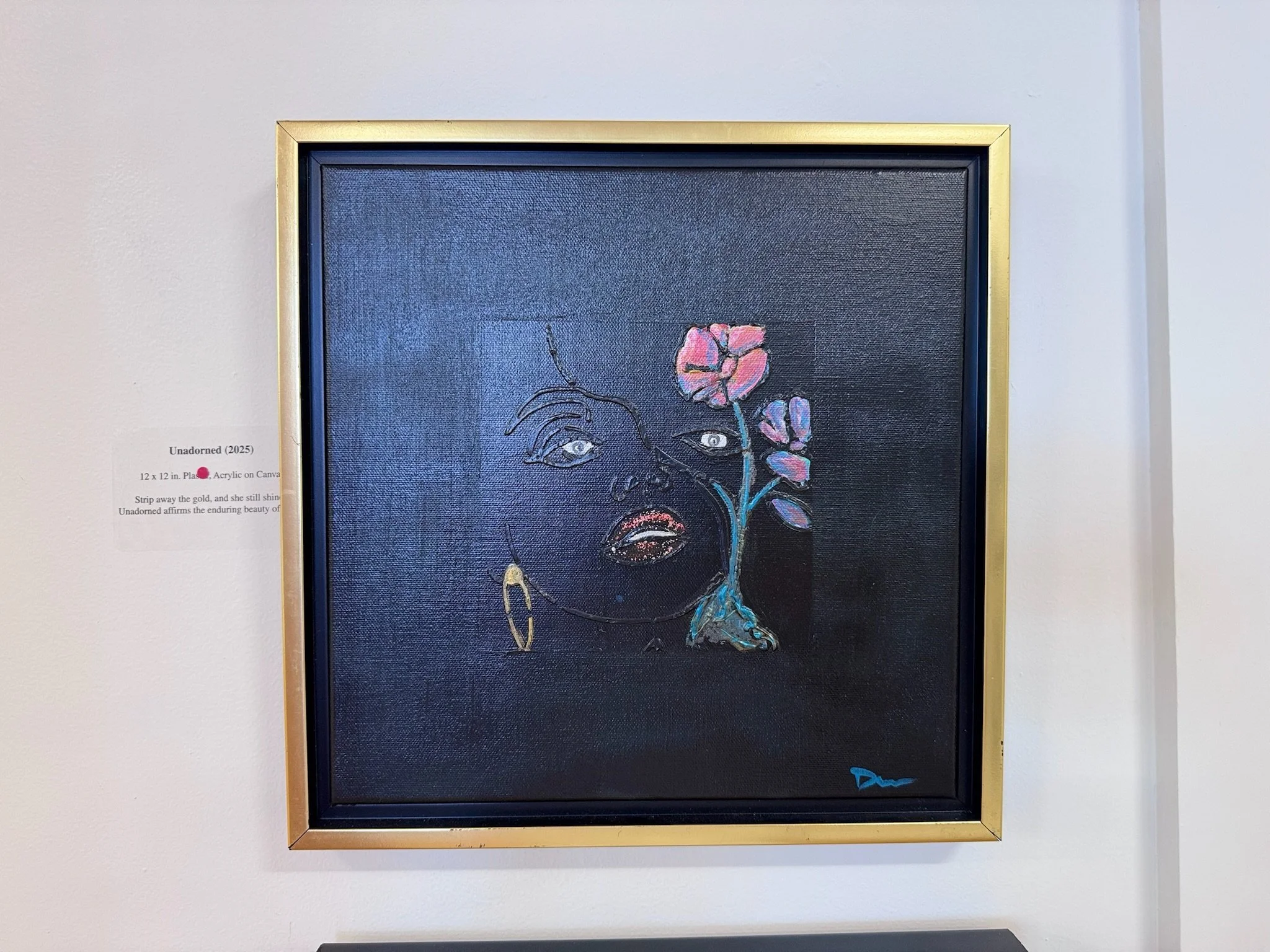An abstract portrait of a woman on a black canvas, with her face outlined in white, a colorful flowering vine growing from her eye, and a gold earring. Framed in gold, displayed on a white wall.