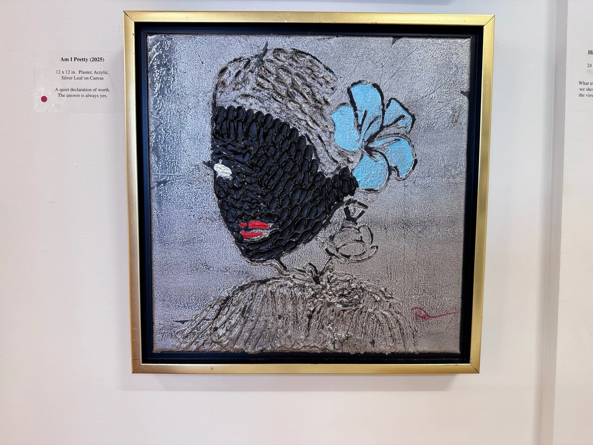 Mixed media art piece of a woman's face with black textured skin, red lips, a gray background, a blue flower in her hair, and a sketchy outline of her clothing, framed in a gold frame.
