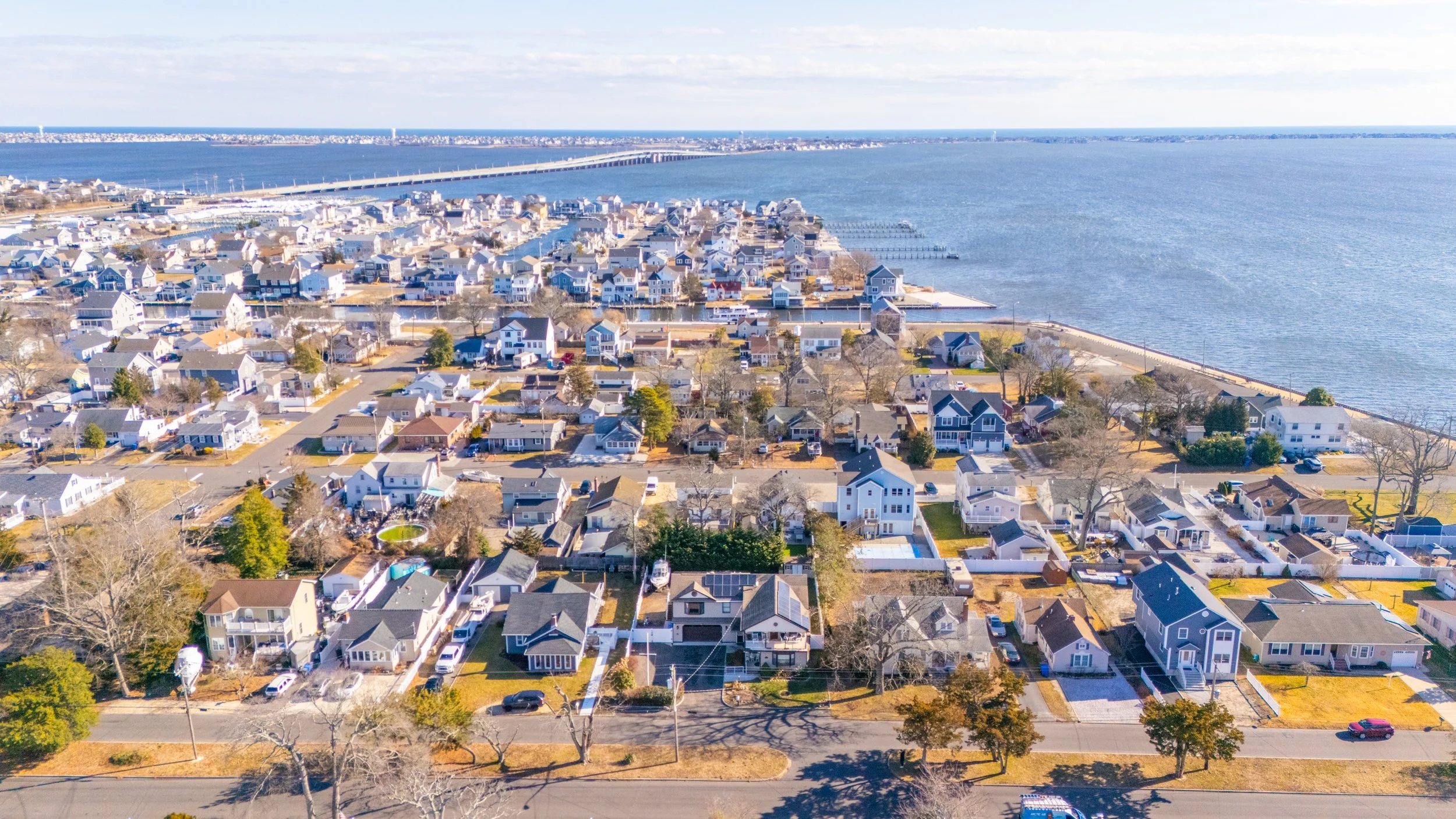 Aerial view of a coastal residential neighborhood with houses, streets, trees, and a body of water in the background, featuring a bridge and waterfront docks.