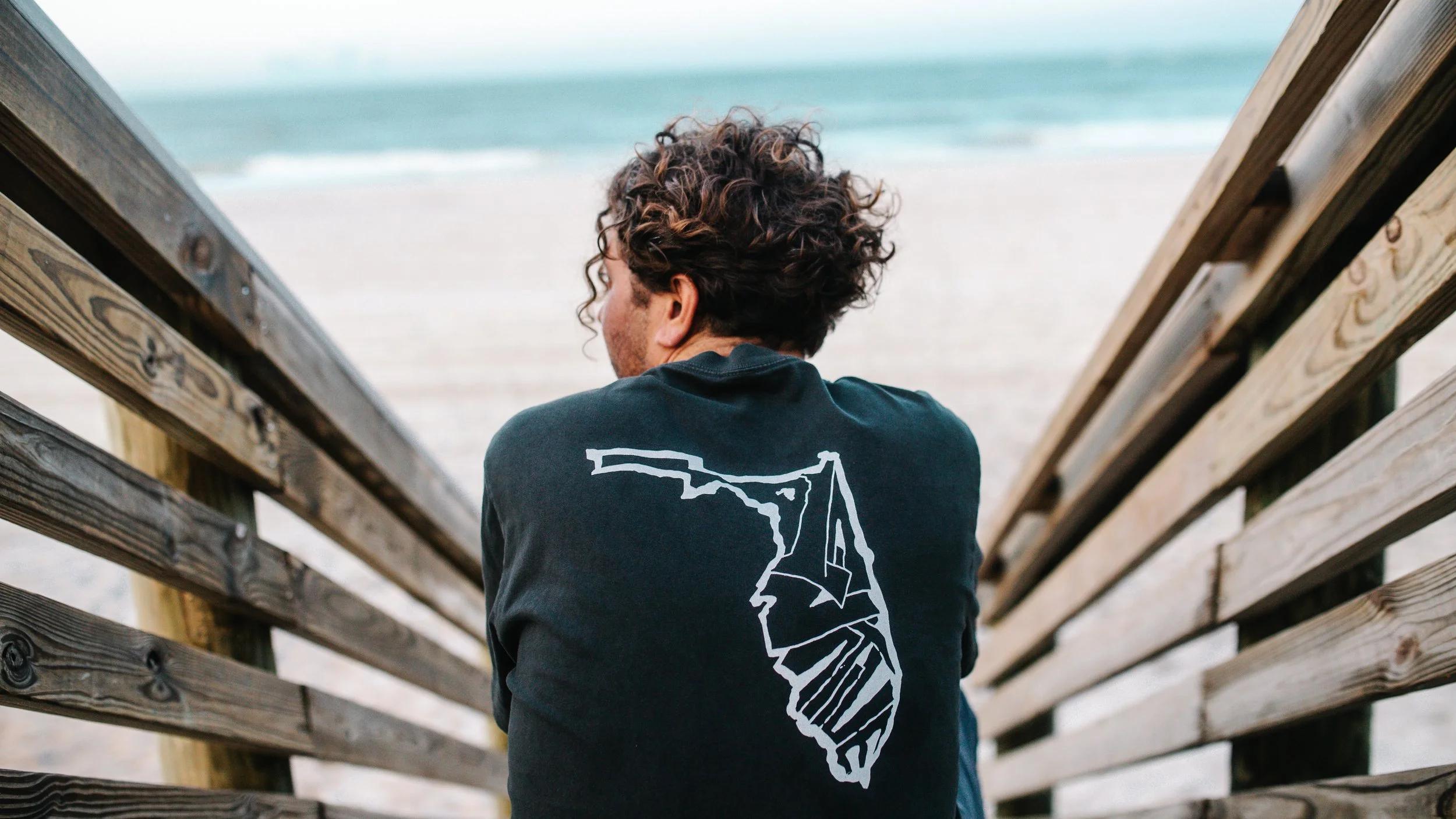 A person with curly hair sitting on a wooden pier, facing the ocean, wearing a black sweatshirt with a white outline of the state of California on the back.