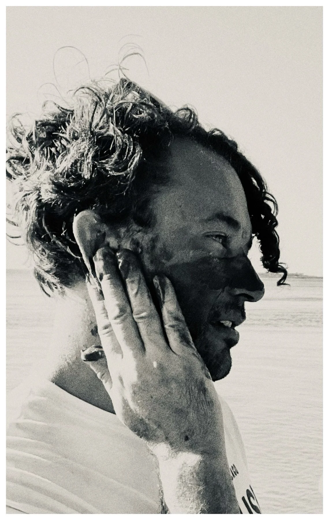 Black and white photo of a man with curly hair, smiling, touching his ear with his hand, near a body of water.