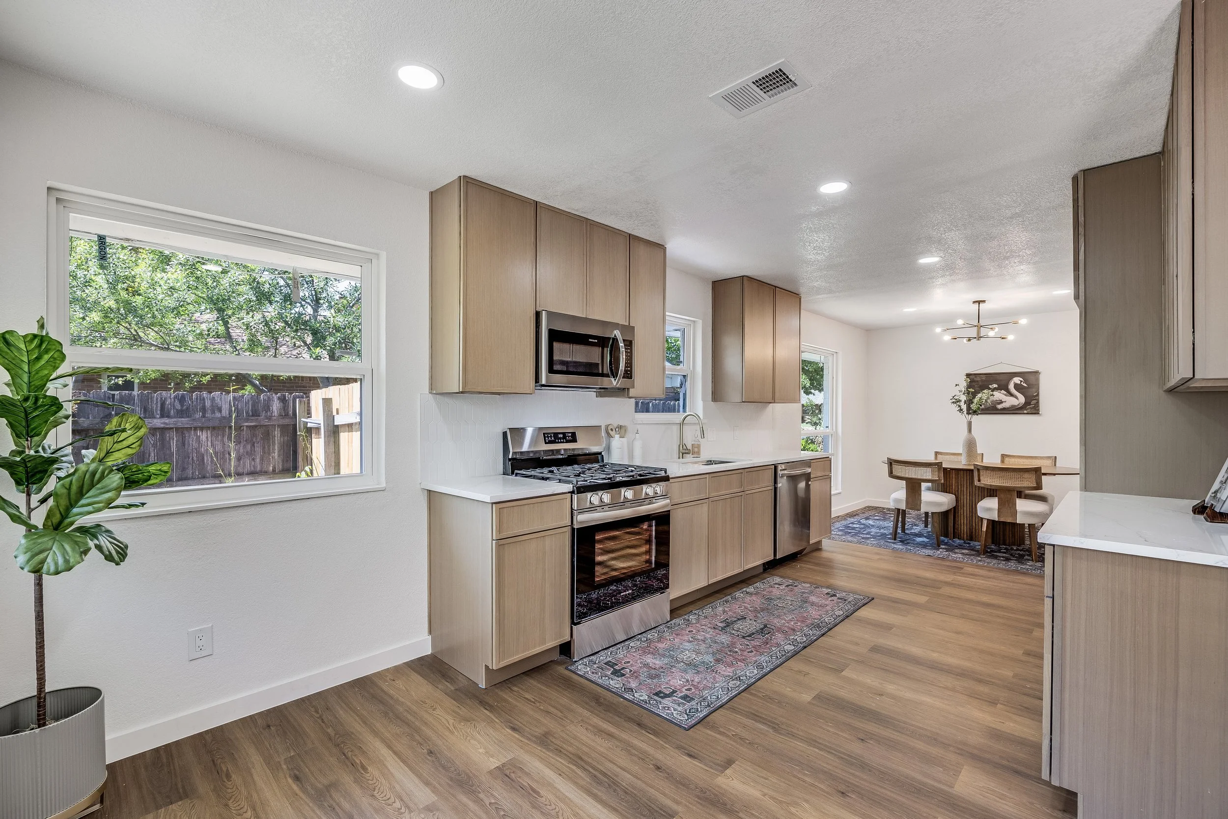 Modern kitchen with light wood cabinets, stainless steel stove and microwave, white quartz countertops, a small dining area with a round table and four chairs, and a large window bringing in natural light.