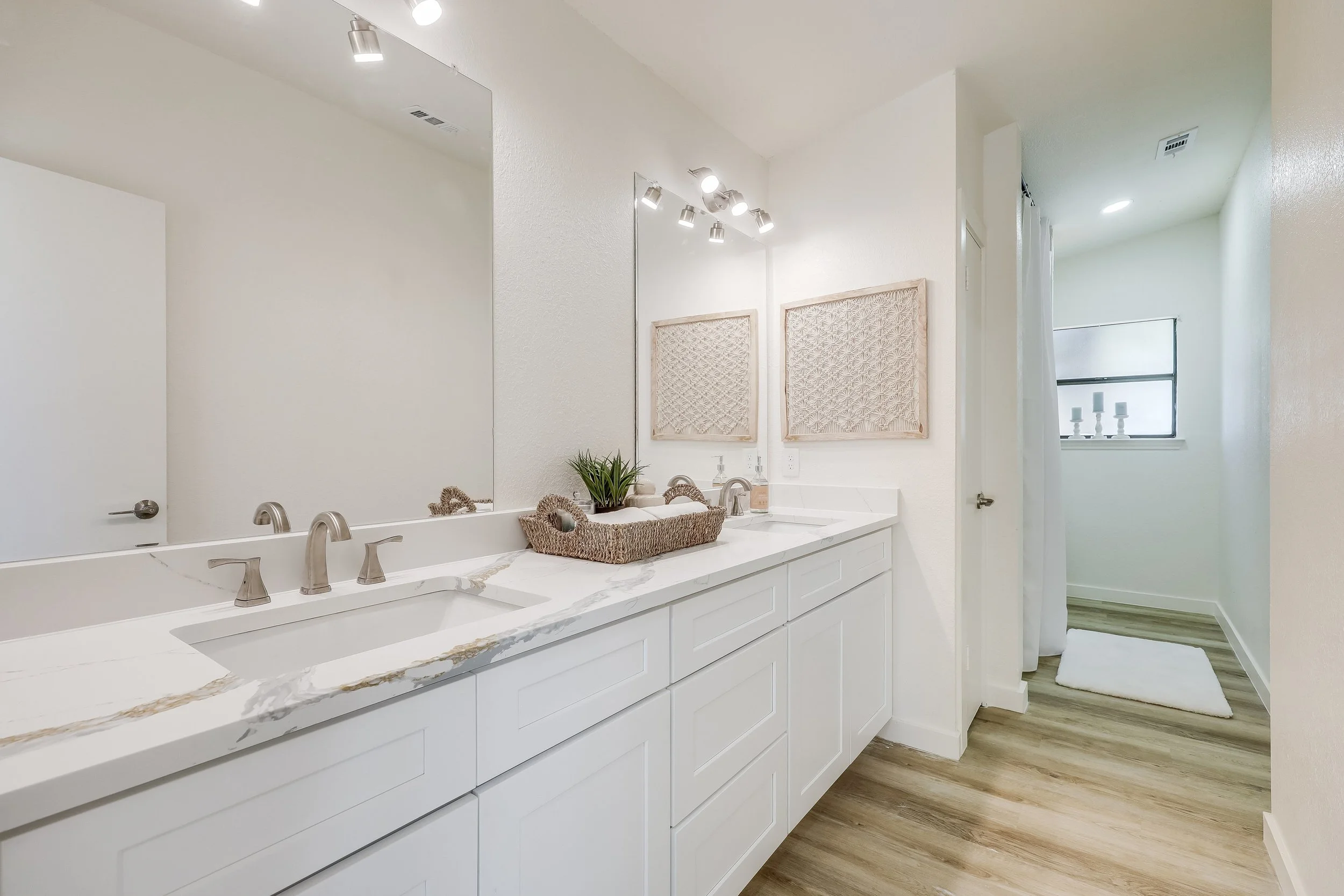 Bright bathroom with white double vanity, marble countertop, large mirror, wall-mounted lights, decorative wall art, small potted plant, basket with toiletries, window with white curtains, light wood flooring.