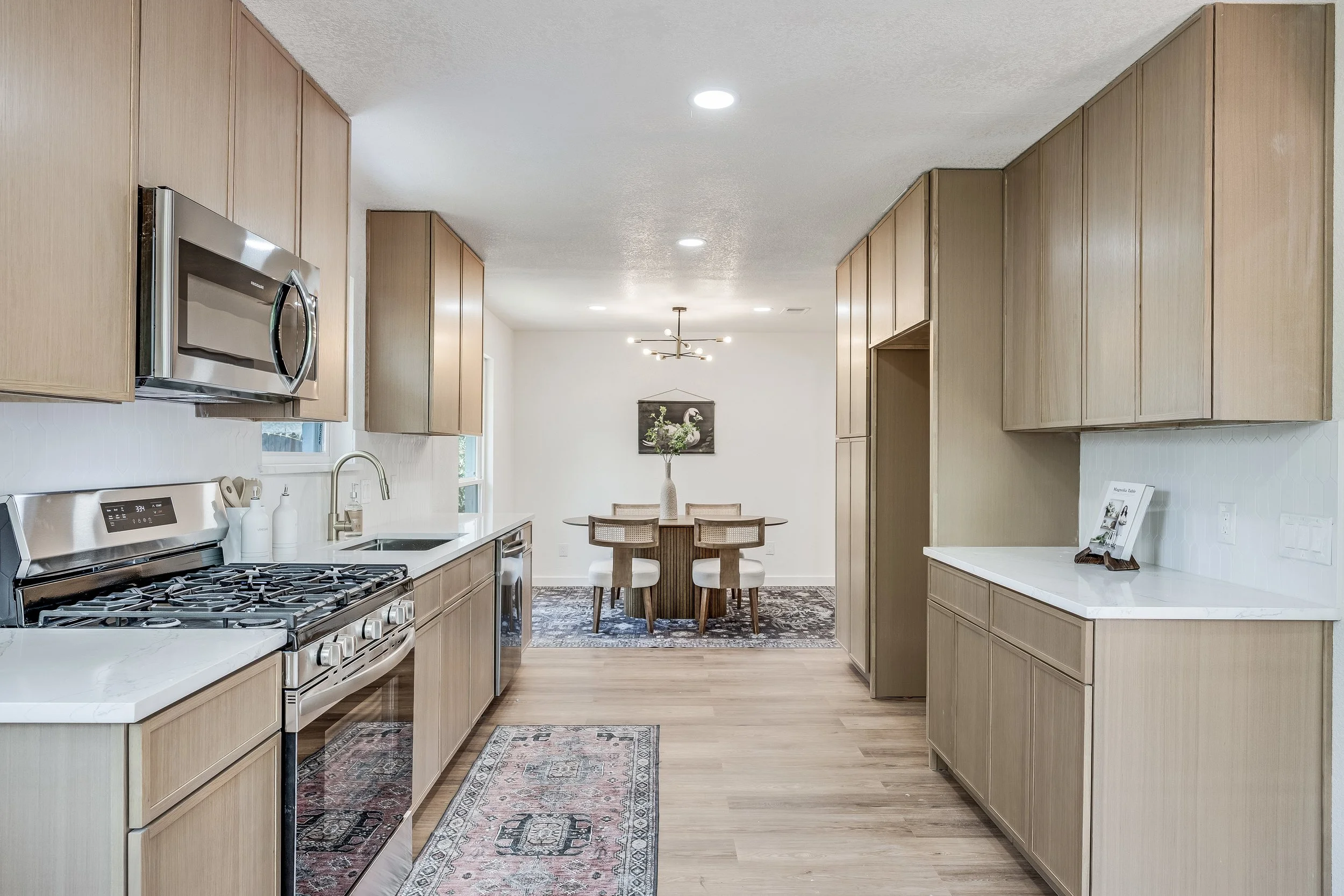 Kitchen with wooden cabinets, stainless steel microwave and stove, white countertops, hardwood floors, and a dining area with a round table, four chairs, a vase with flowers, and a framed picture of a swan hanging on the white wall.