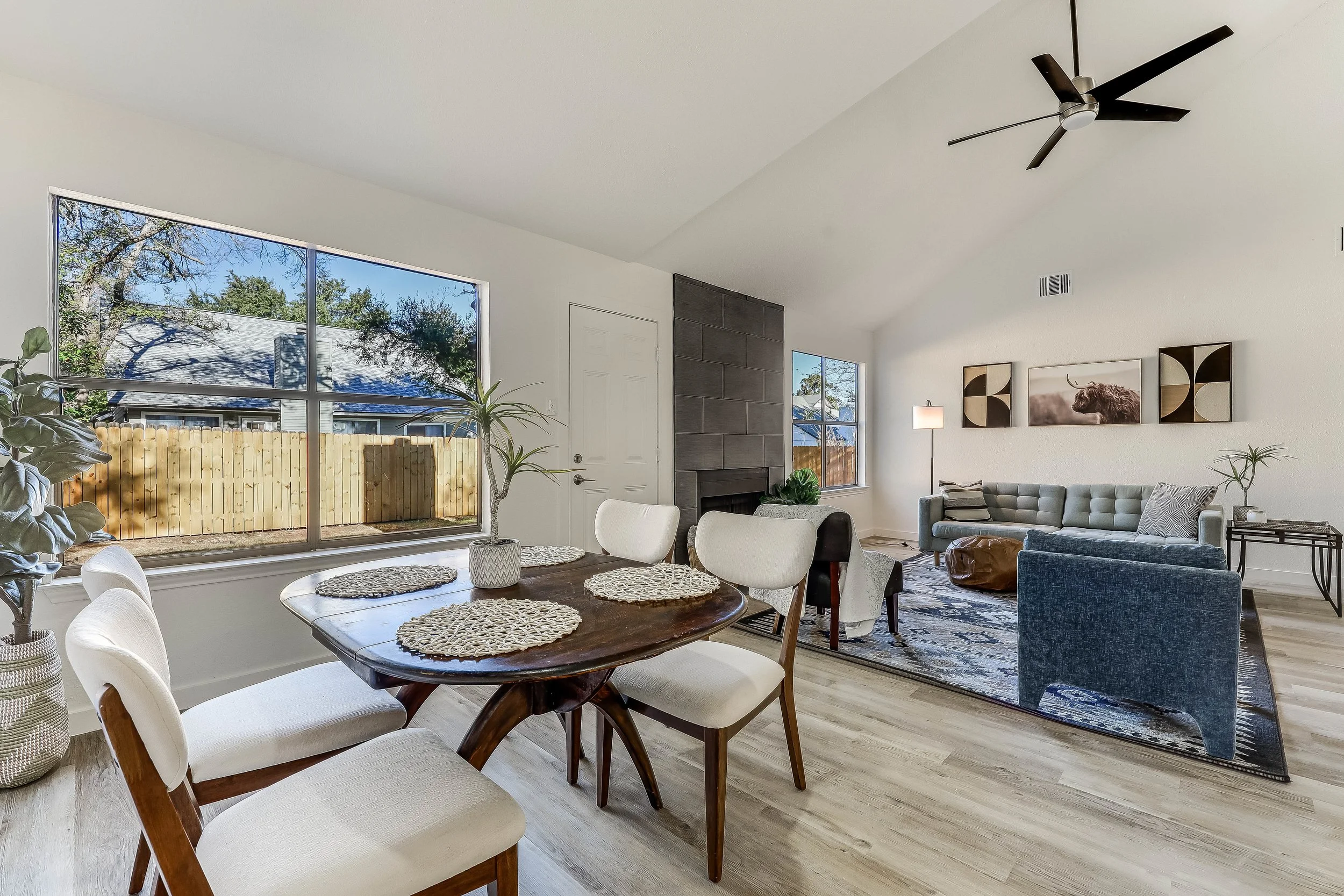 Open living and dining room with large windows, white walls, wooden floors, and modern furniture, including a dining table with chairs, a sofa, armchair, and artwork on the wall.