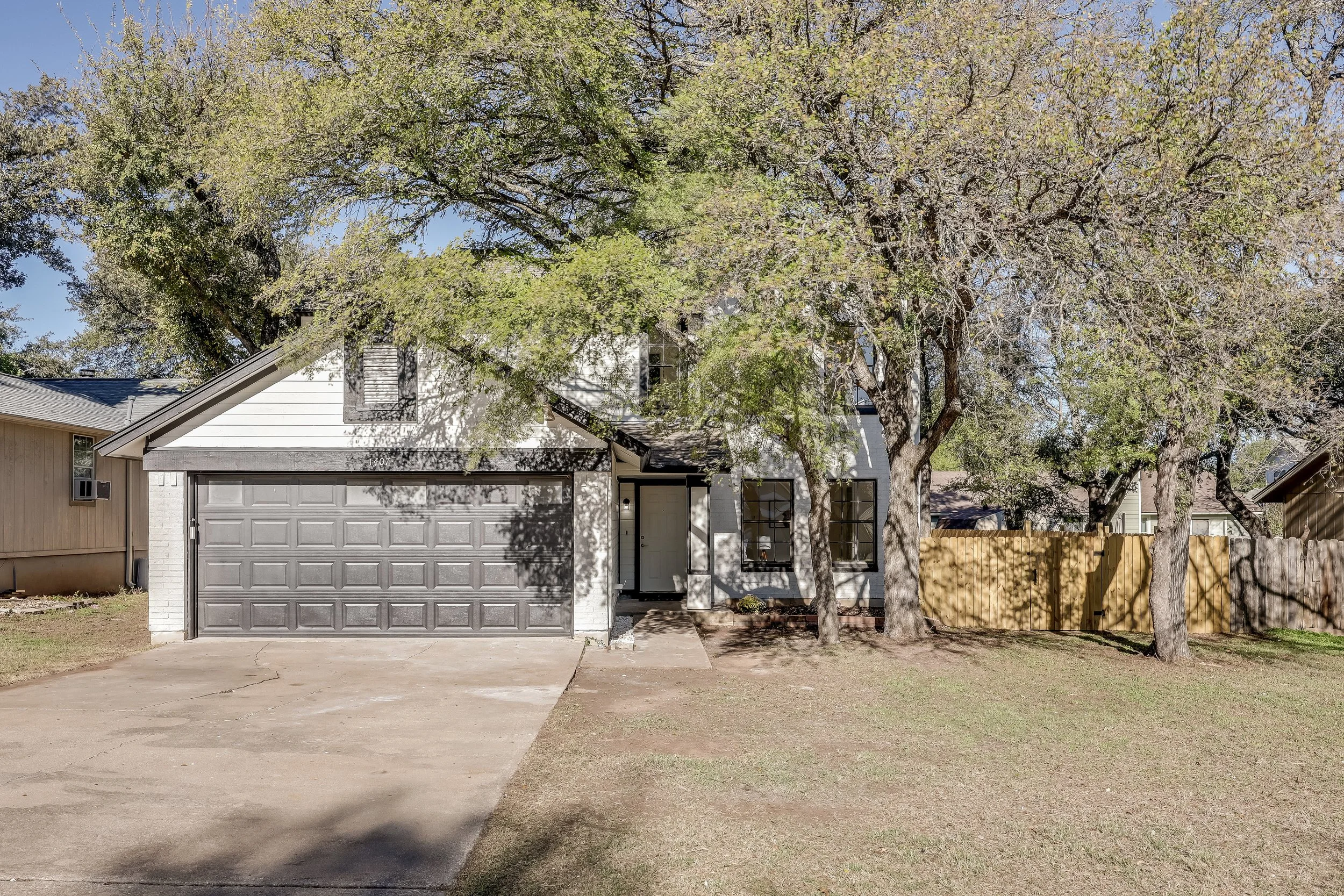 A two-story residential house with a white exterior, gray garage door, front porch, and large trees in the yard, surrounded by a wooden fence on the right side.