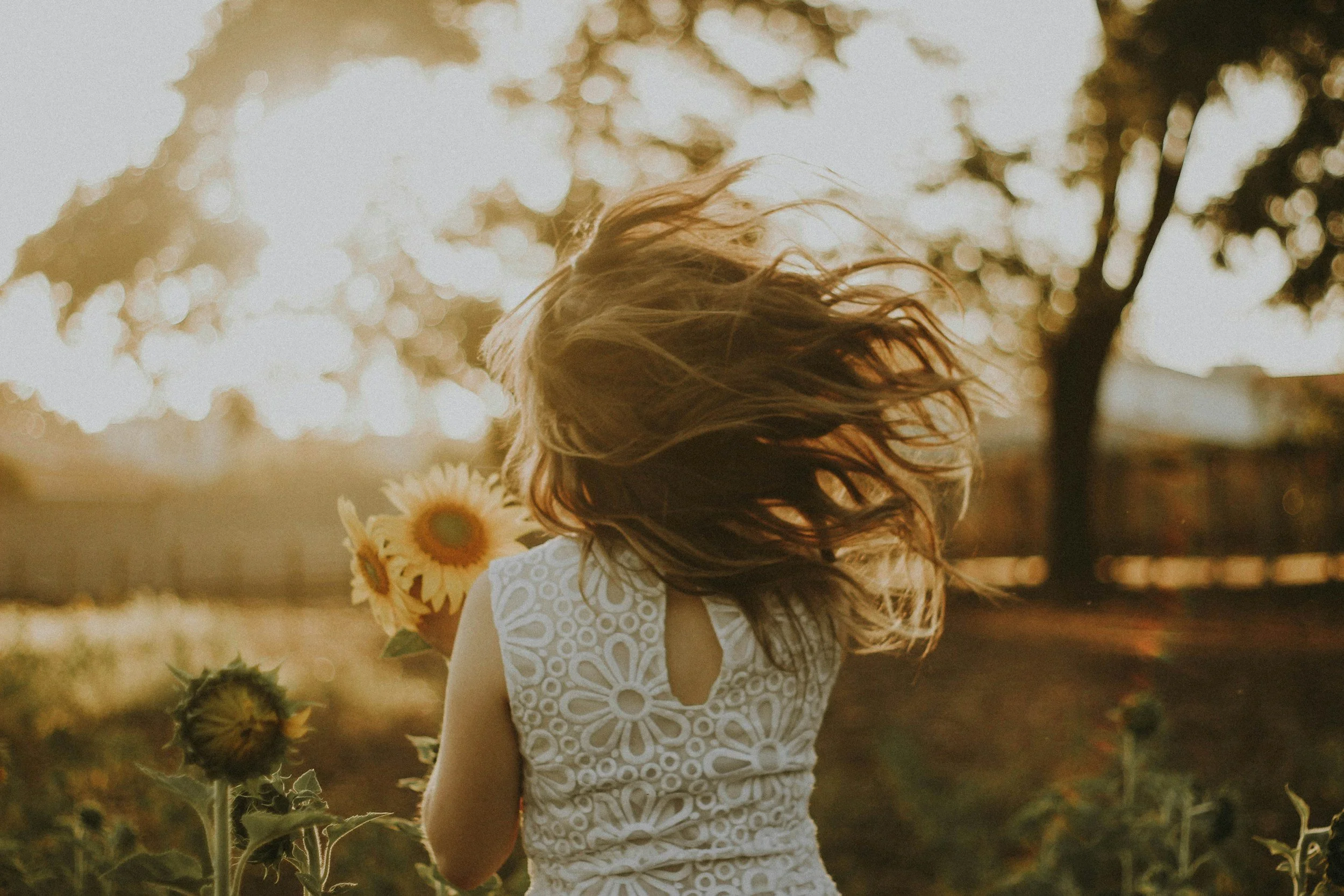 A woman outdoors with her hair blowing in the wind, holding a sunflower, wearing a white lace dress, with trees and sunlight in the background.