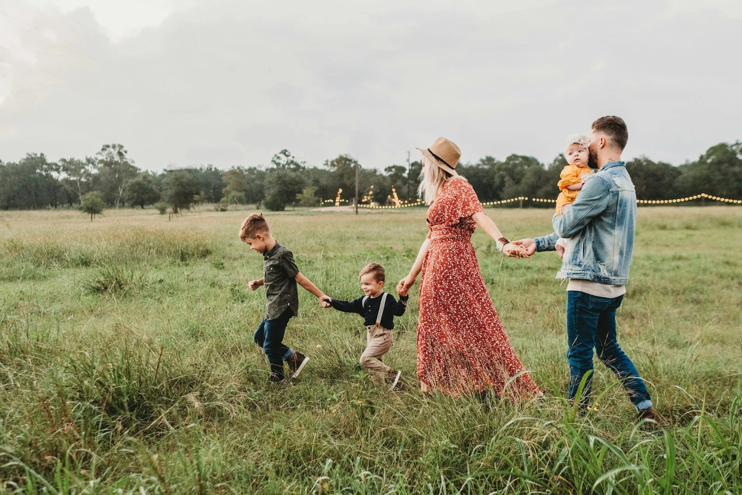 Family walking together in a grassy field, holding hands, with a cloudy sky and string lights in the background.