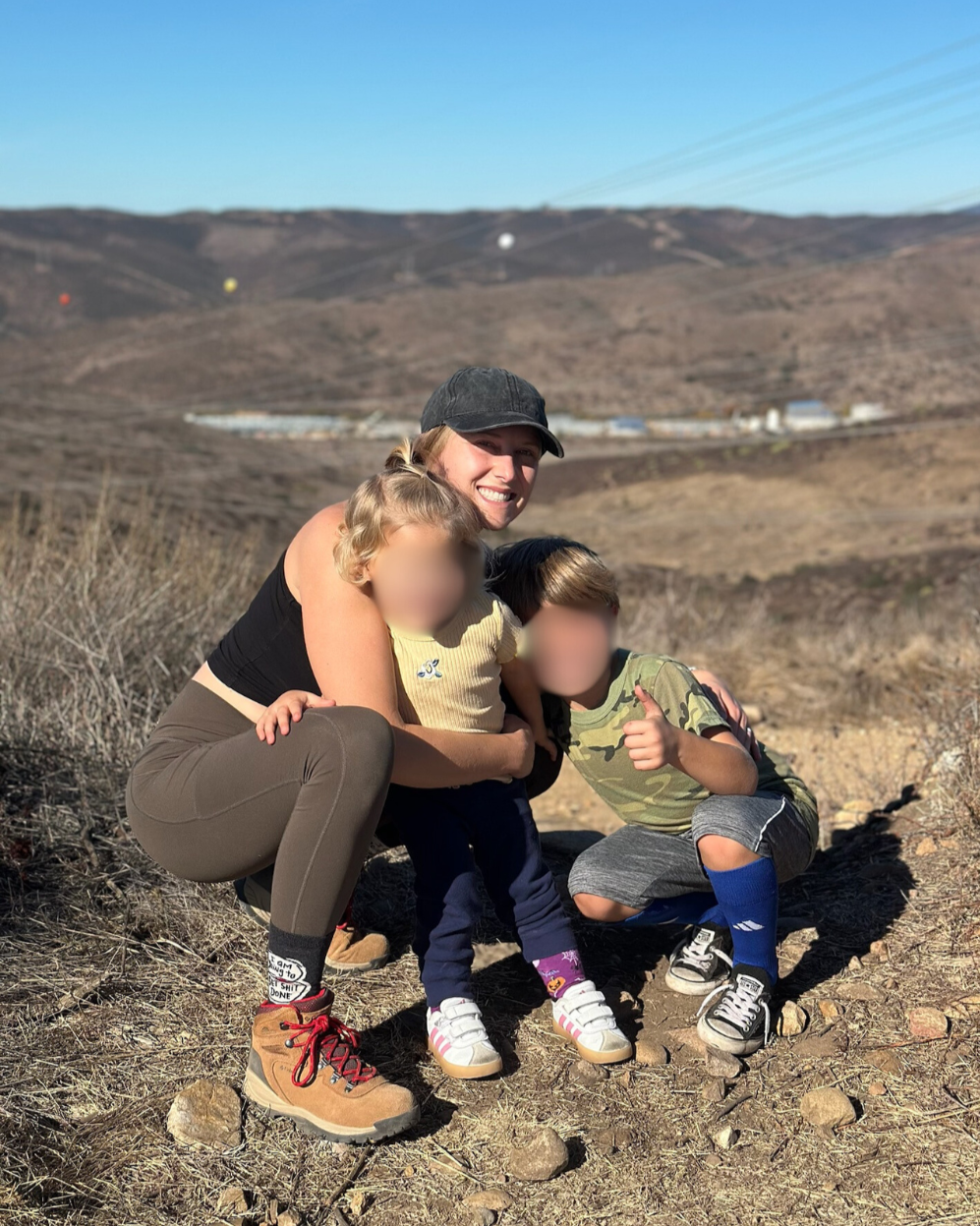 A woman smiling with two children, one girl and one boy, outdoors on a rocky terrain with a mountainous landscape in the background. All are dressed casually for a hike or outdoor activity.