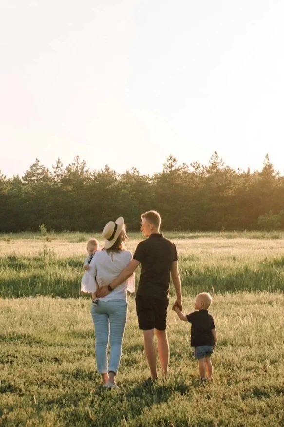 A family of four walking through a grassy field at sunset, holding hands, with trees in the background.