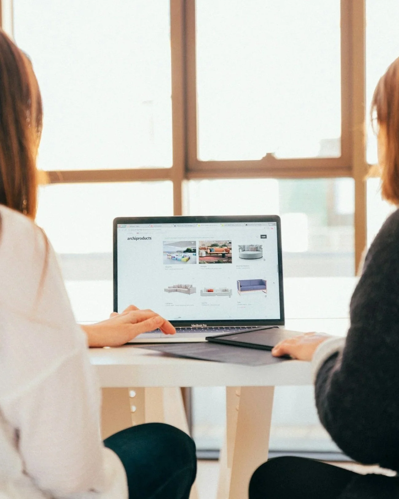 Two women sitting at a table looking at a laptop screen displaying furniture images on a website.