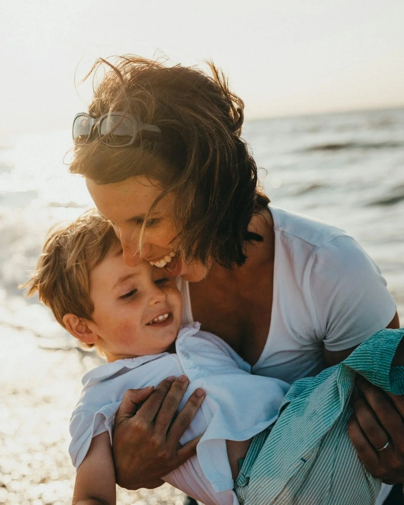 A woman and a young boy are playing and hugging on the beach near the ocean during sunset.