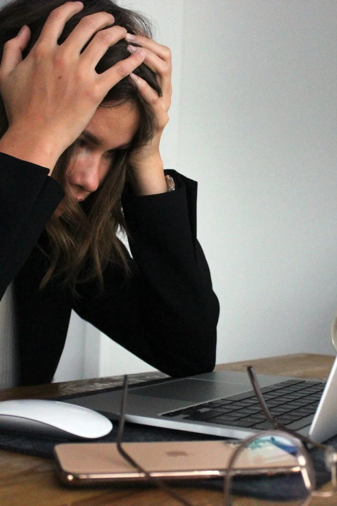 woman in black shirt holding her head in frustration while seated at a desk with a laptop, glasses, and a smartphone.