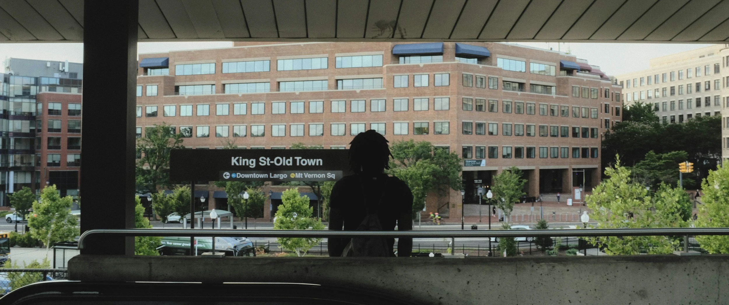 Person silhouetted in front of King St-Old Town metro station sign with brick office buildings and greenery in the background.