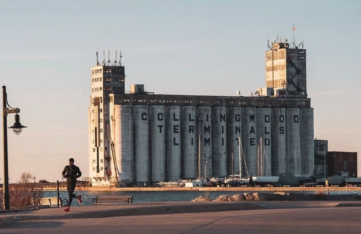 A person jogging near a large industrial building labeled "Collingwood Terminals Limited," located by the waterfront. The building has multiple silos and a tower with a flag. The scene is set during sunset, casting a warm glow.