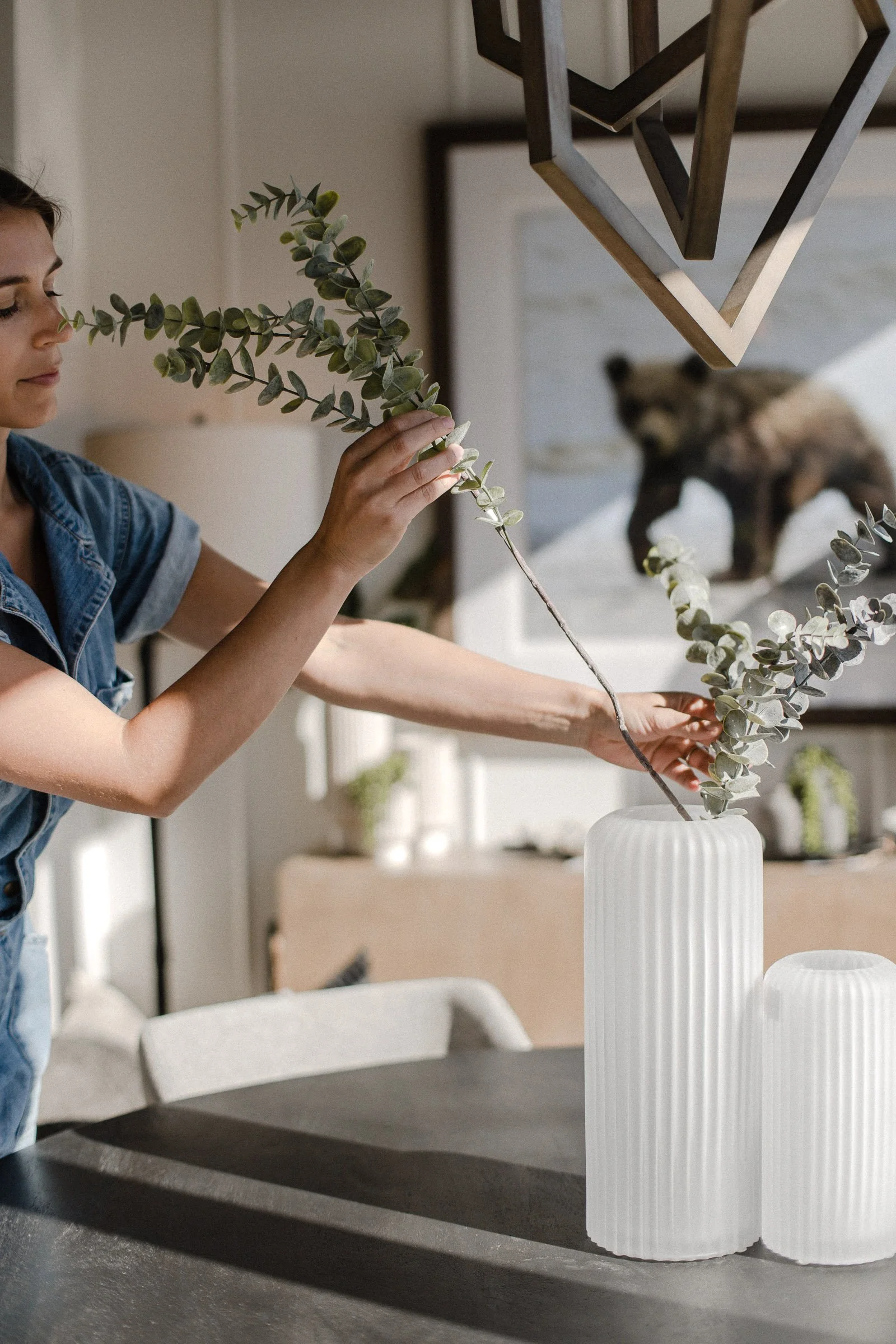 Person arranging eucalyptus branches in a white vase on a table with a framed bear picture in the background.