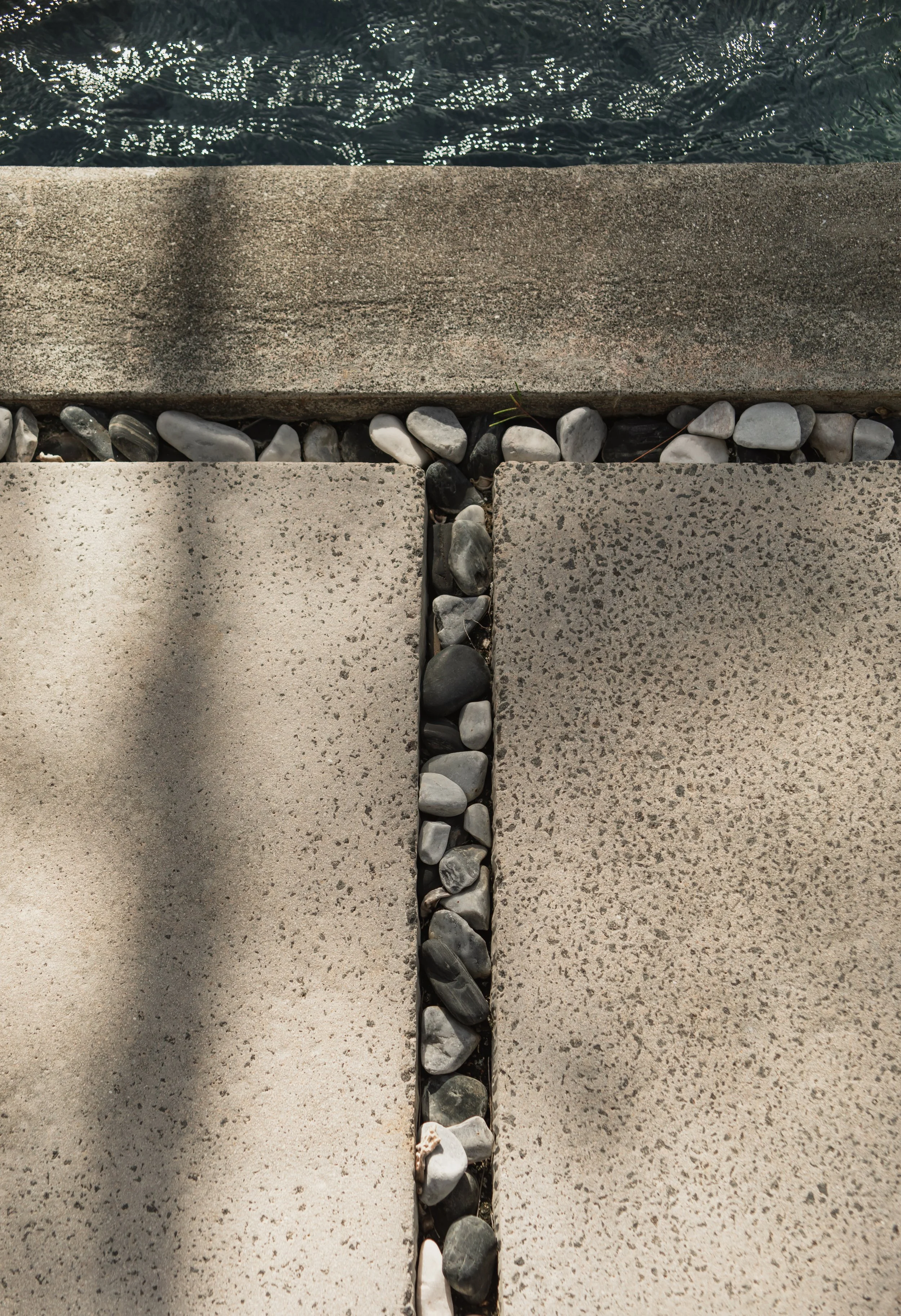 Close-up of a concrete paving with a gap filled with small black and white pebbles, next to a stone edge and a water surface in the background.