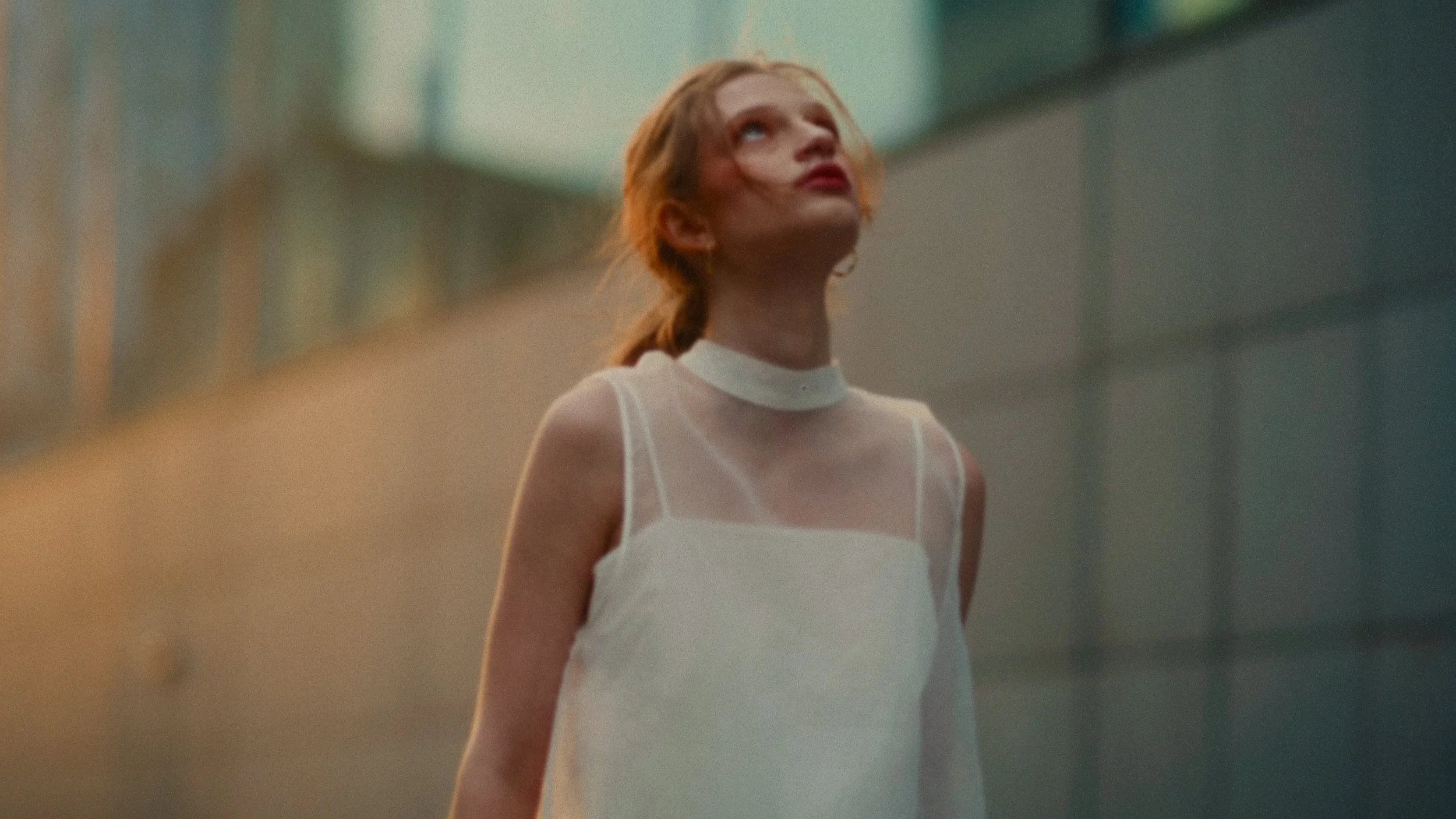 A woman with red hair, wearing a white sleeveless top, standing outdoors in an urban setting with glass buildings in the background, looking upwards.