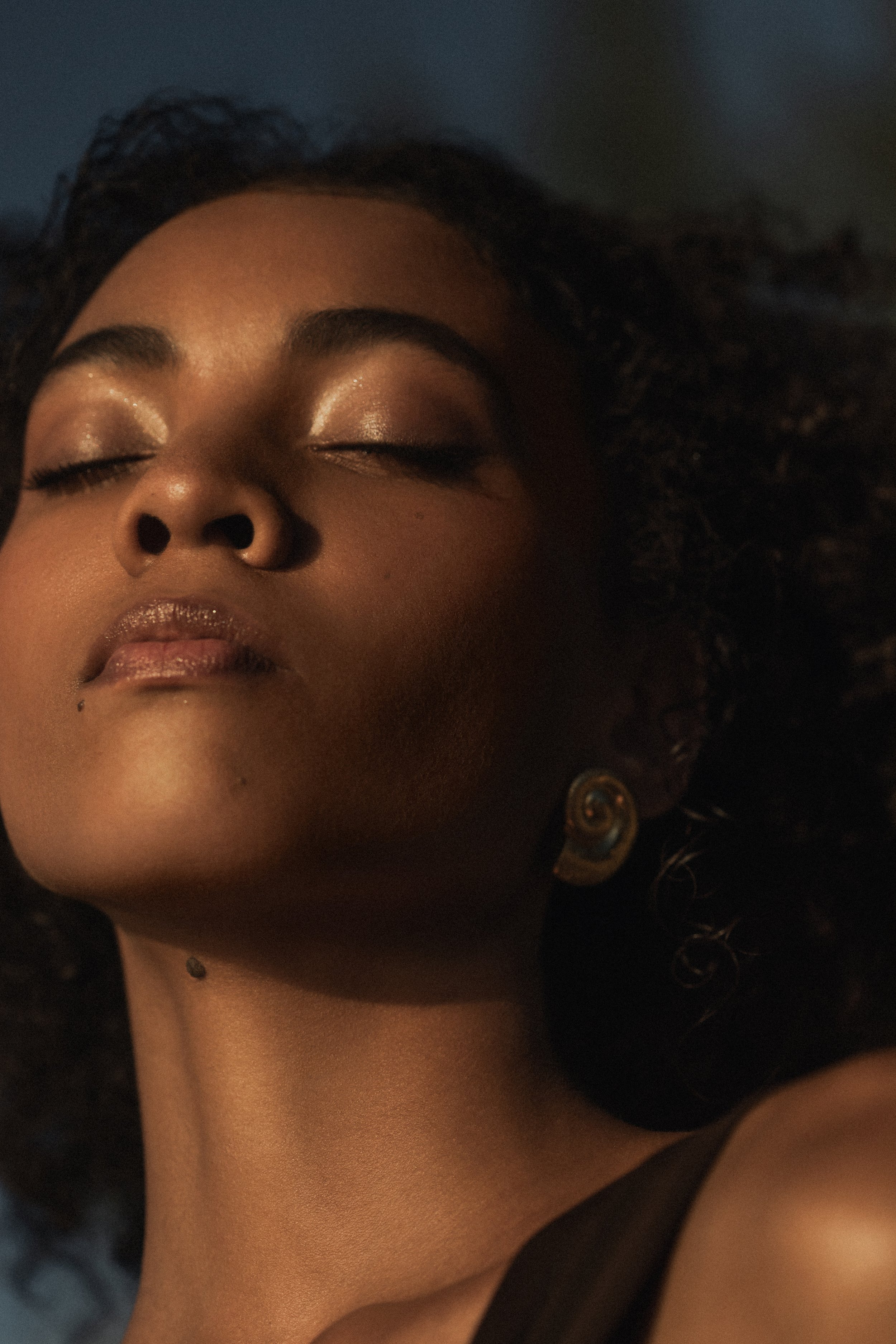 Close-up of a woman's face with her eyes closed, makeup, and earrings, lying on a dark surface.