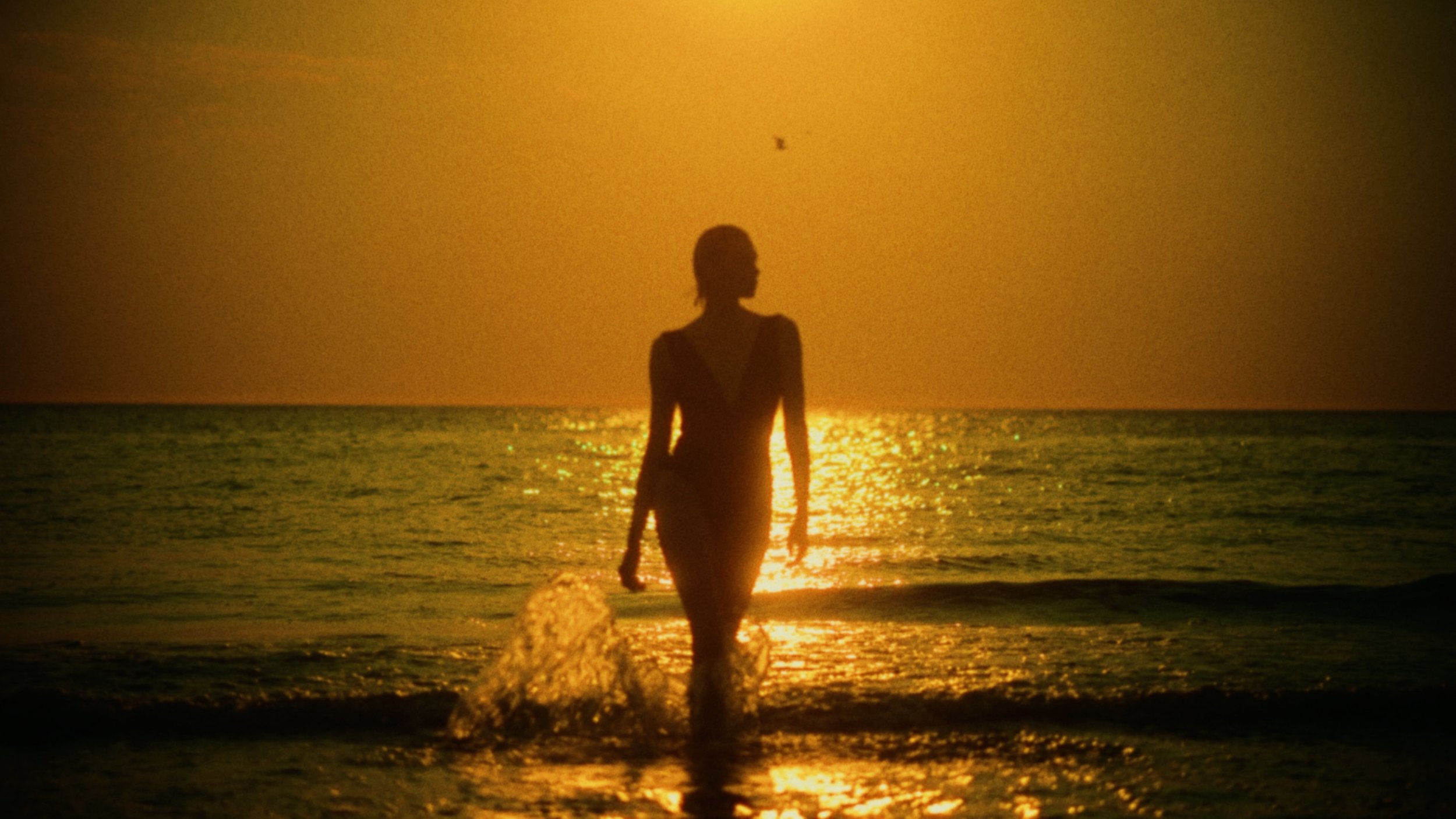 Silhouette of a woman walking in the ocean at sunset, with the sun reflecting on the water and a clear sky.