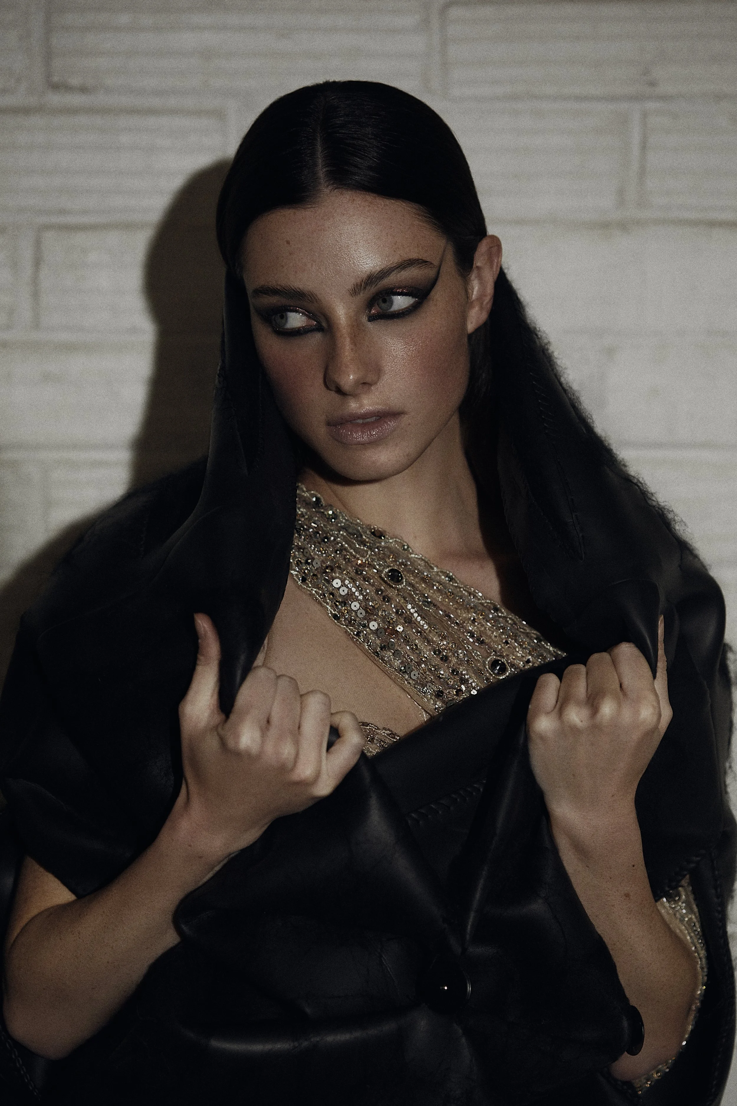 A woman with long black hair and striking makeup wears a sparkling top and black leather jacket, standing against a white brick wall.