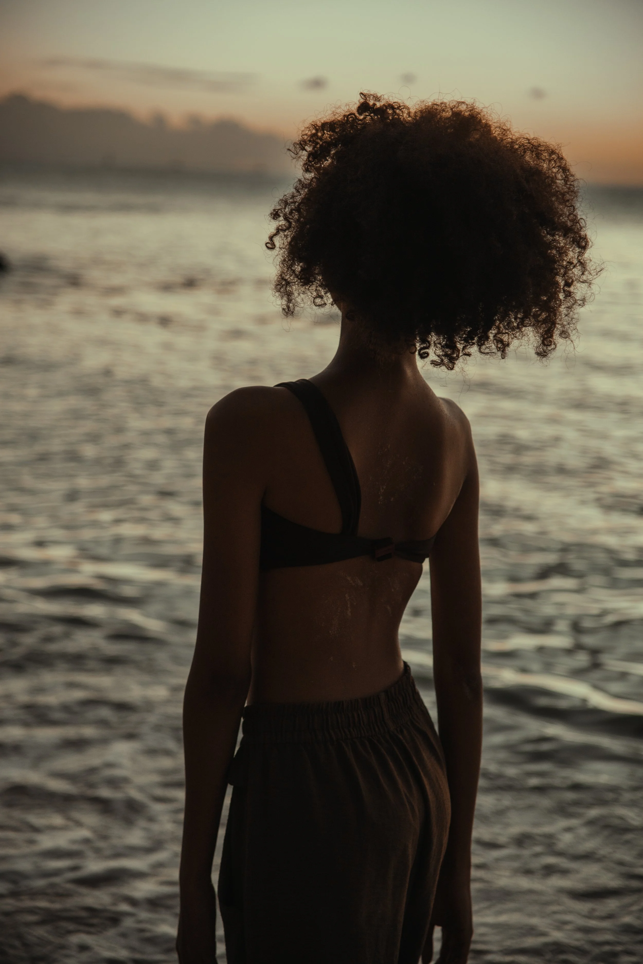 A silhouette of a woman with curly hair standing on a beach during sunset, facing the water.