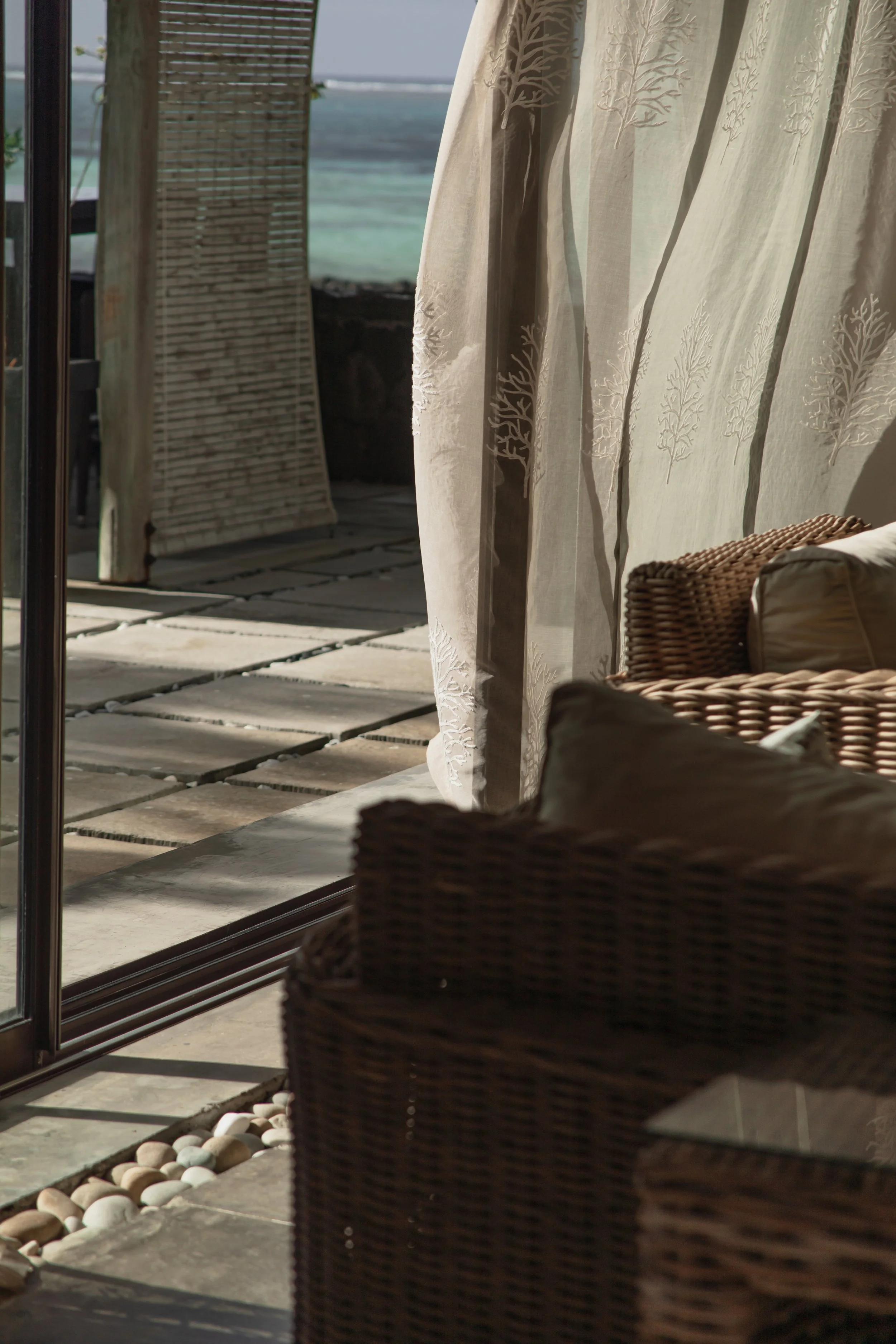 View of a beach scene through a window with curtains, featuring wicker furniture and a pathway with white stones.