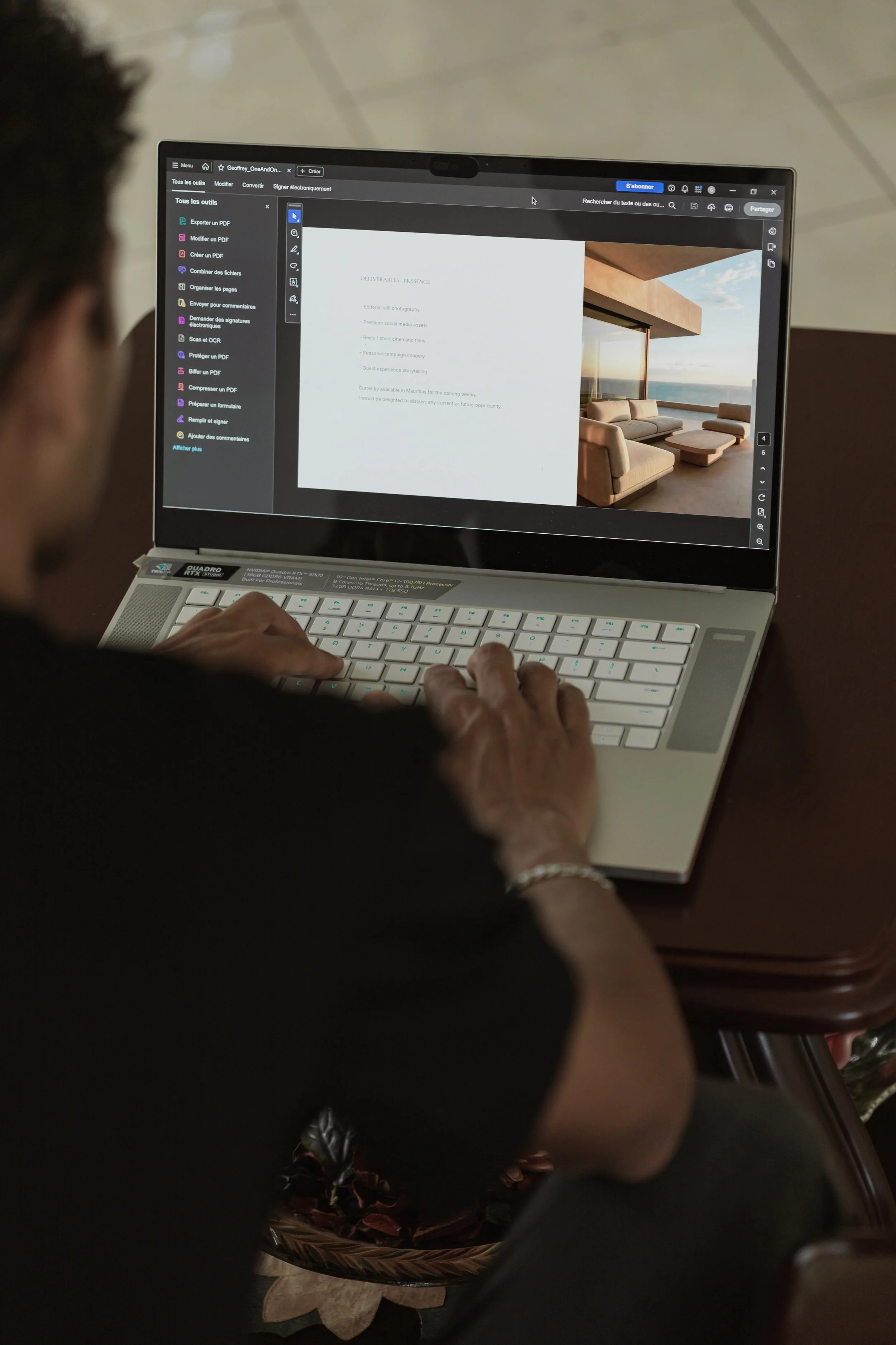 A person using a laptop on a wooden table, with a potted plant nearby. The laptop screen displays a document with text and a background image of a modern living room with large windows and a sunset view.