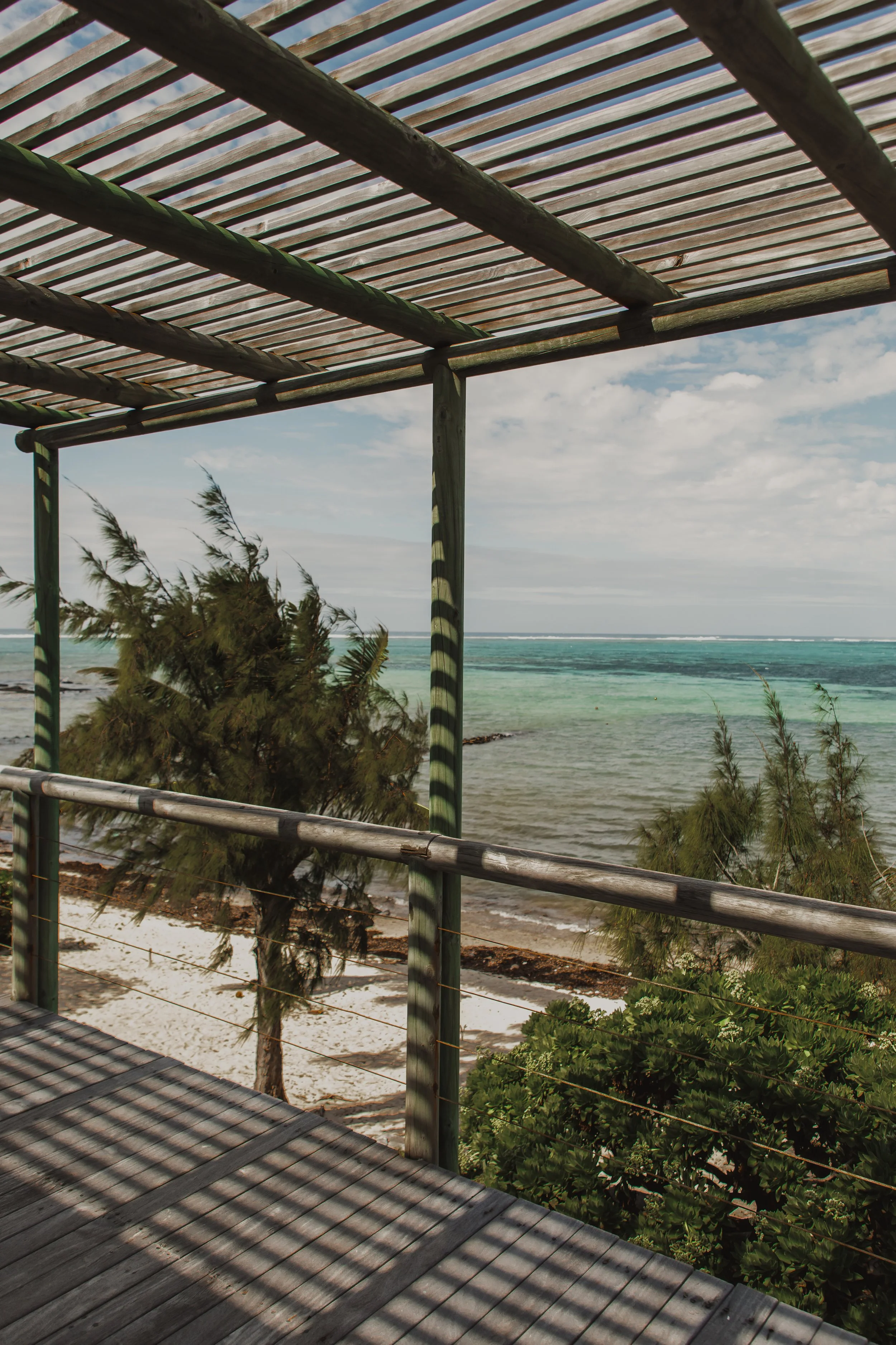 A view from a wooden deck or platform overlooking the beach with seagrass, coastal greenery, and ocean waves, under a partly cloudy sky.