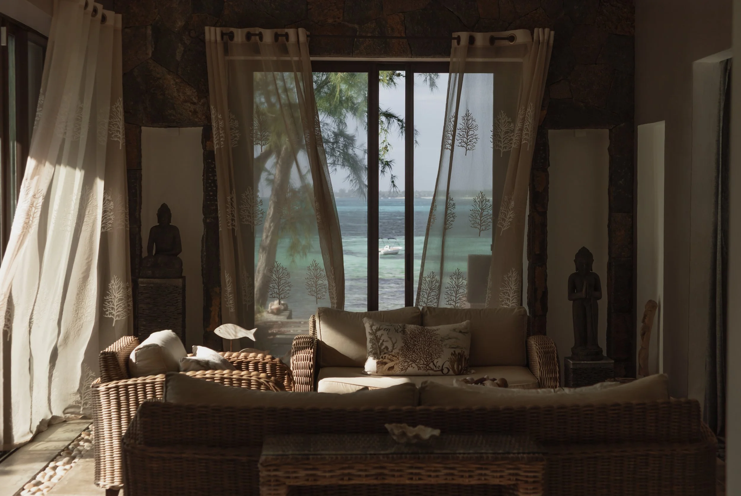 Living room with wicker furniture, cream curtains with tree patterns, statues, and a large window showing a view of the ocean and trees outside.