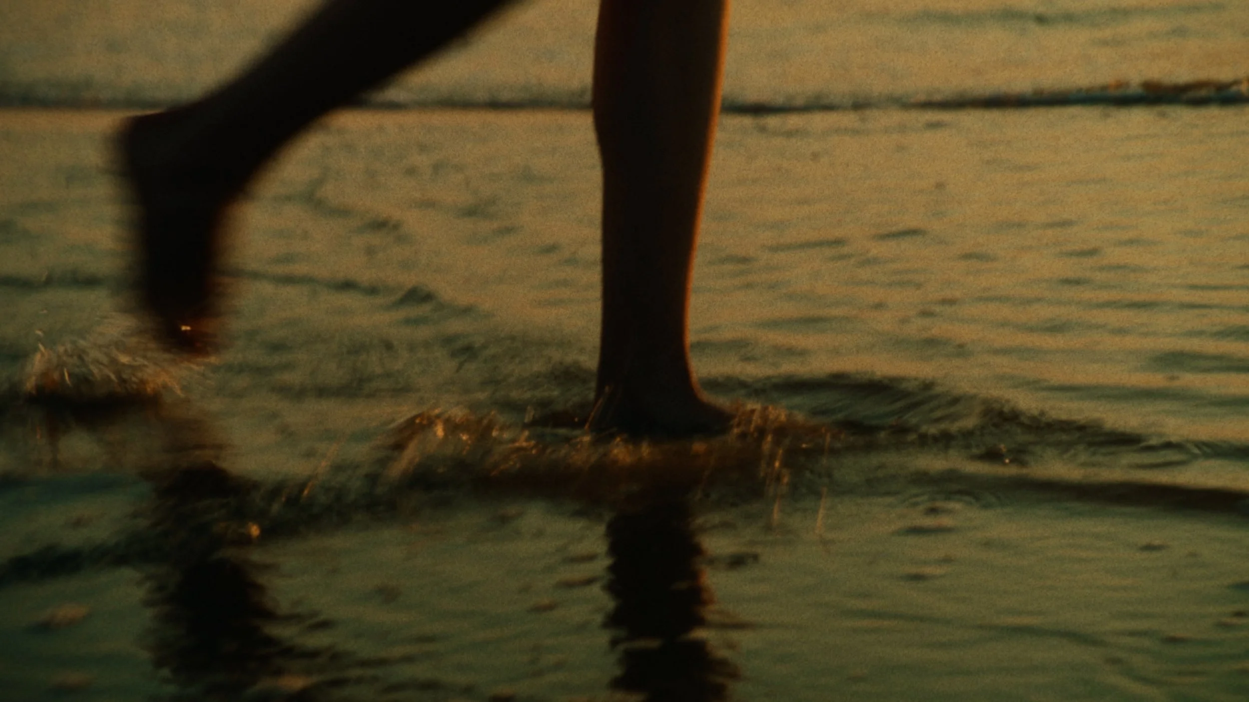 Close-up of a person's legs walking along a calm body of water at sunset, with ripples and reflections on the surface.