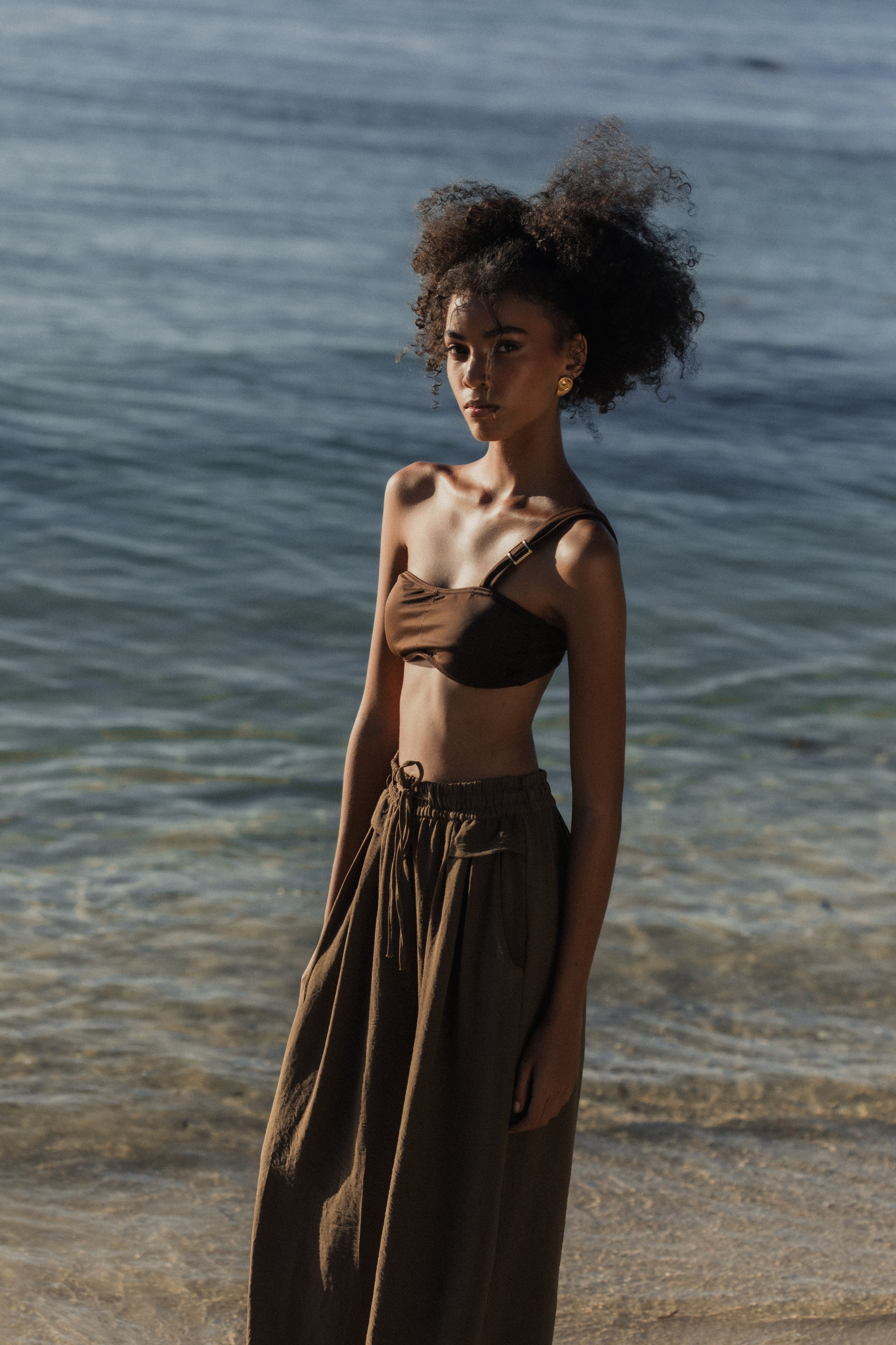 A woman with curly hair in a brown top and brown wide-leg pants standing on a beach near the water.