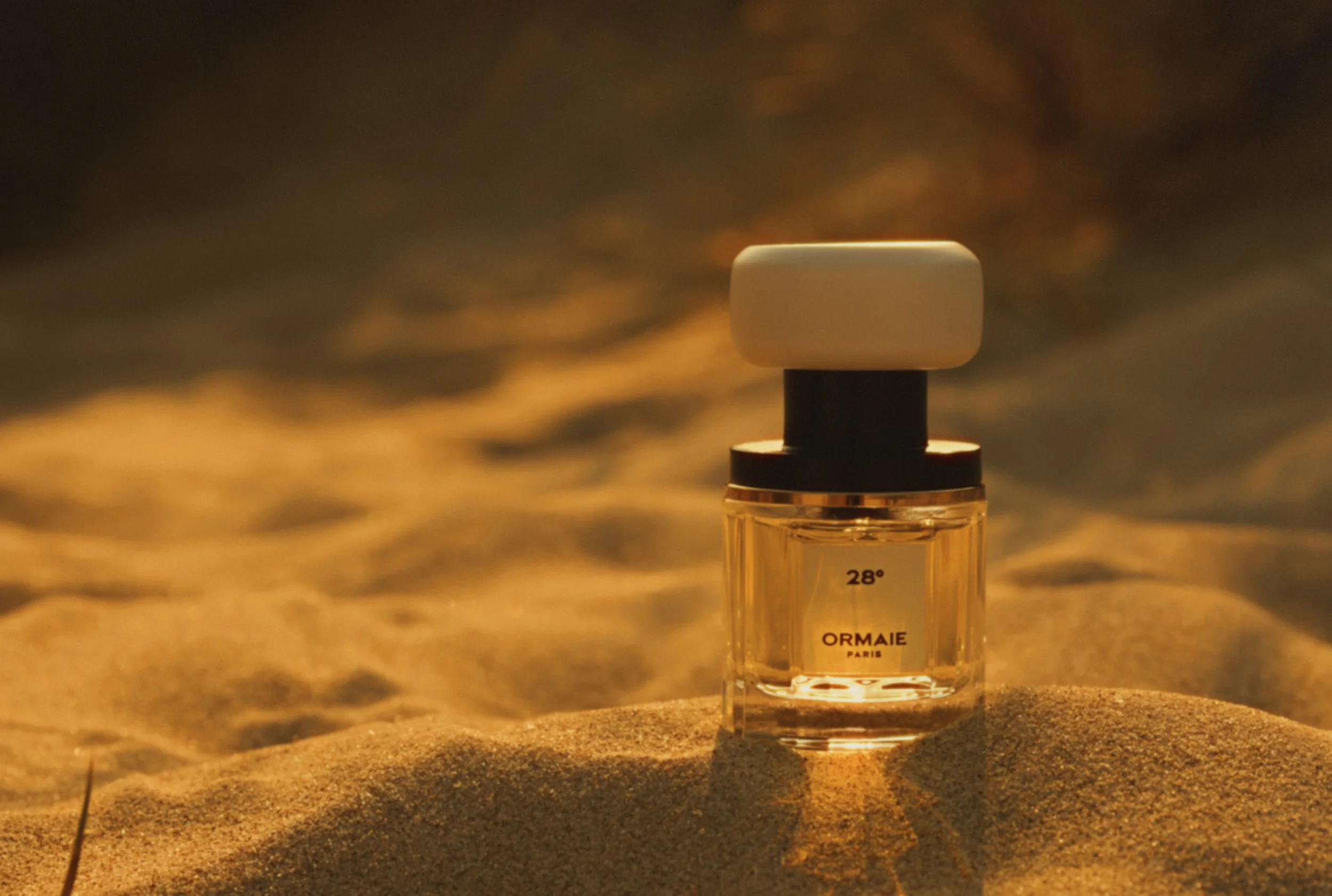 A small perfume bottle with a white cap, placed on sand, with a blurred sunset sky in the background.