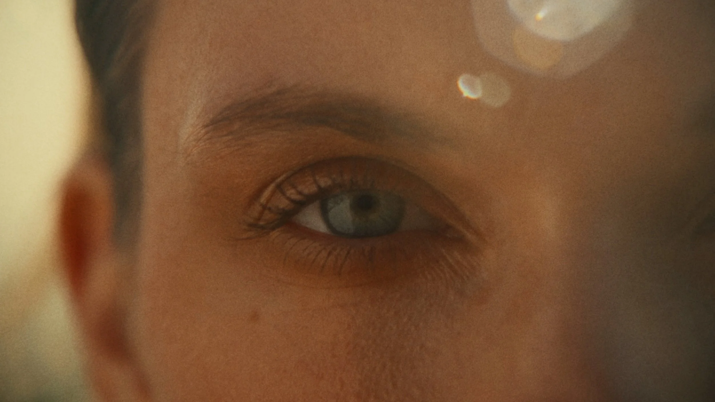 Close-up of a person's eye with light blue iris, eyelashes, and eyebrow, with part of the face and ear visible, and a reflection in the eye.