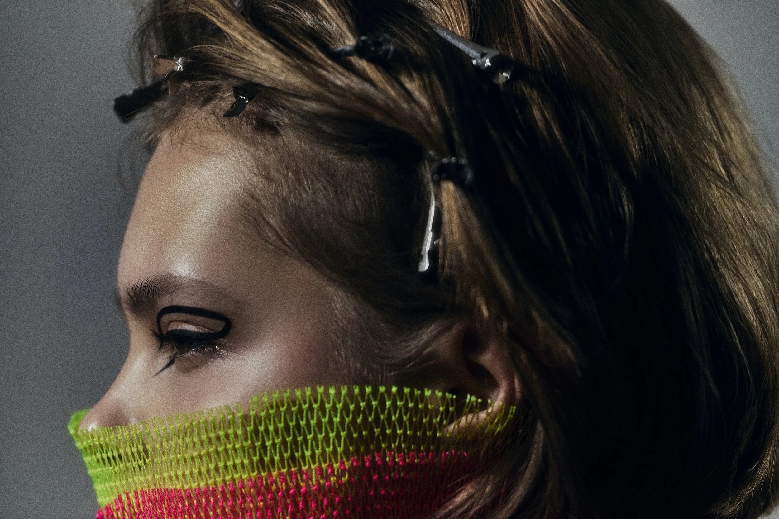 Close-up of a woman's face, showcasing hair with bobby pins, eye makeup with graphic eyeliner, and a bright neon green and pink veil covering her lower face.