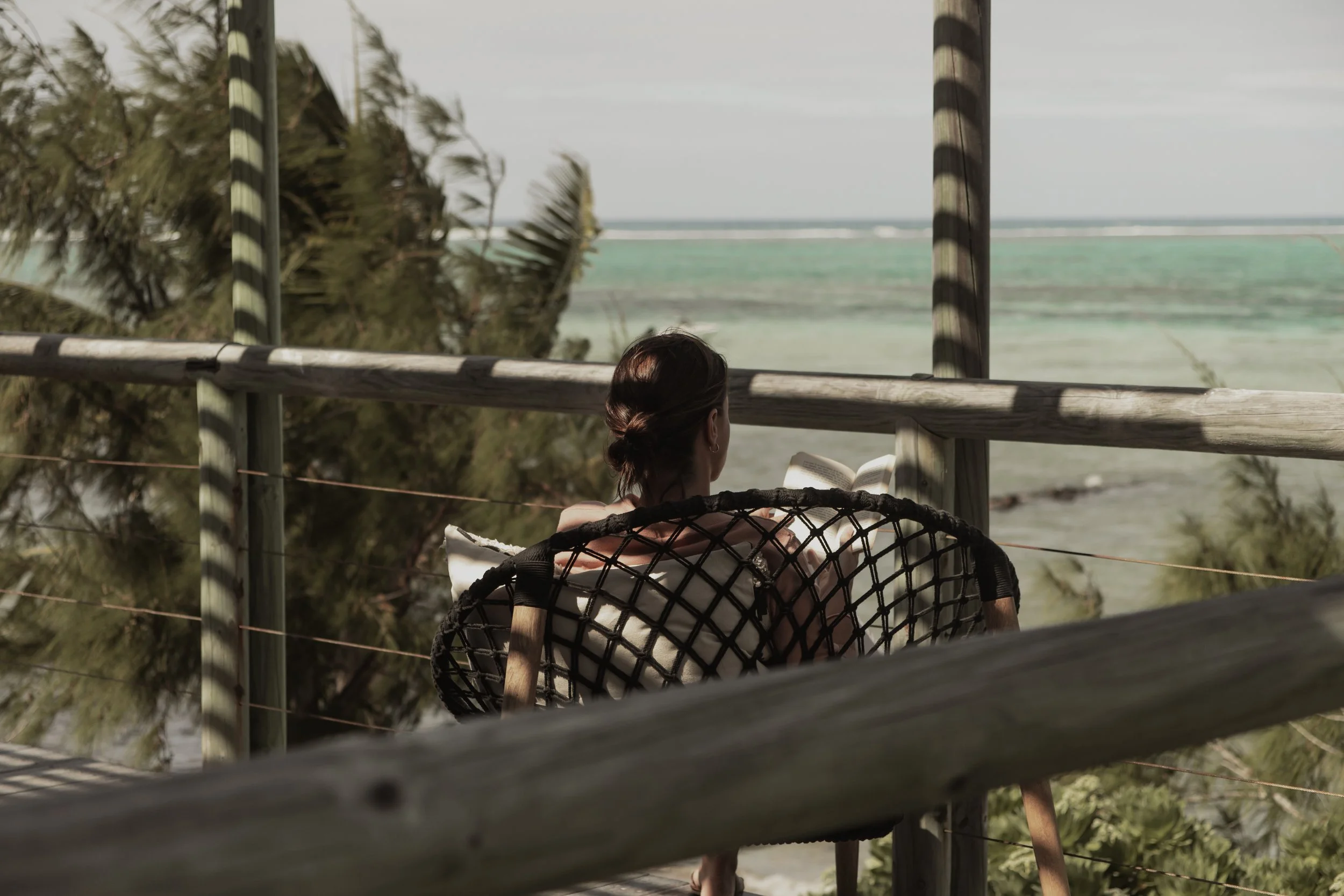 A woman sitting in a chair on a balcony, reading a book, with ocean waves in the background and wind blowing palm trees.