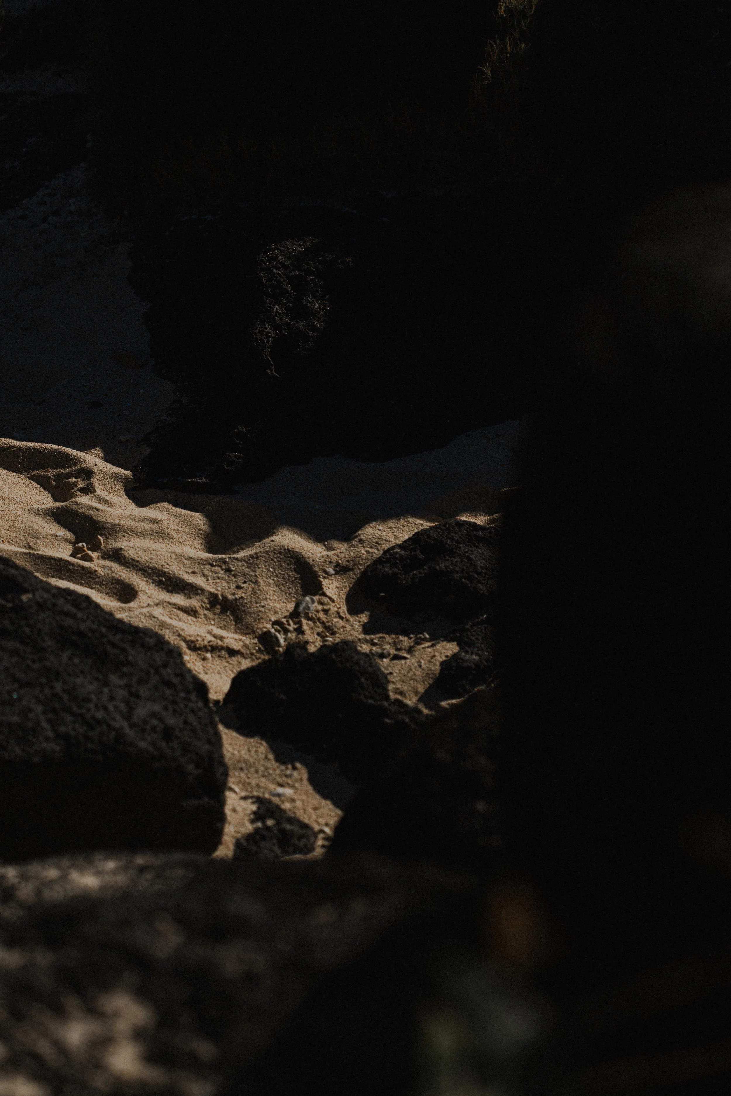 Dark rocks and sand on a beach, with some sunlight illuminating the surface and a shadowed rocky formation in the background.