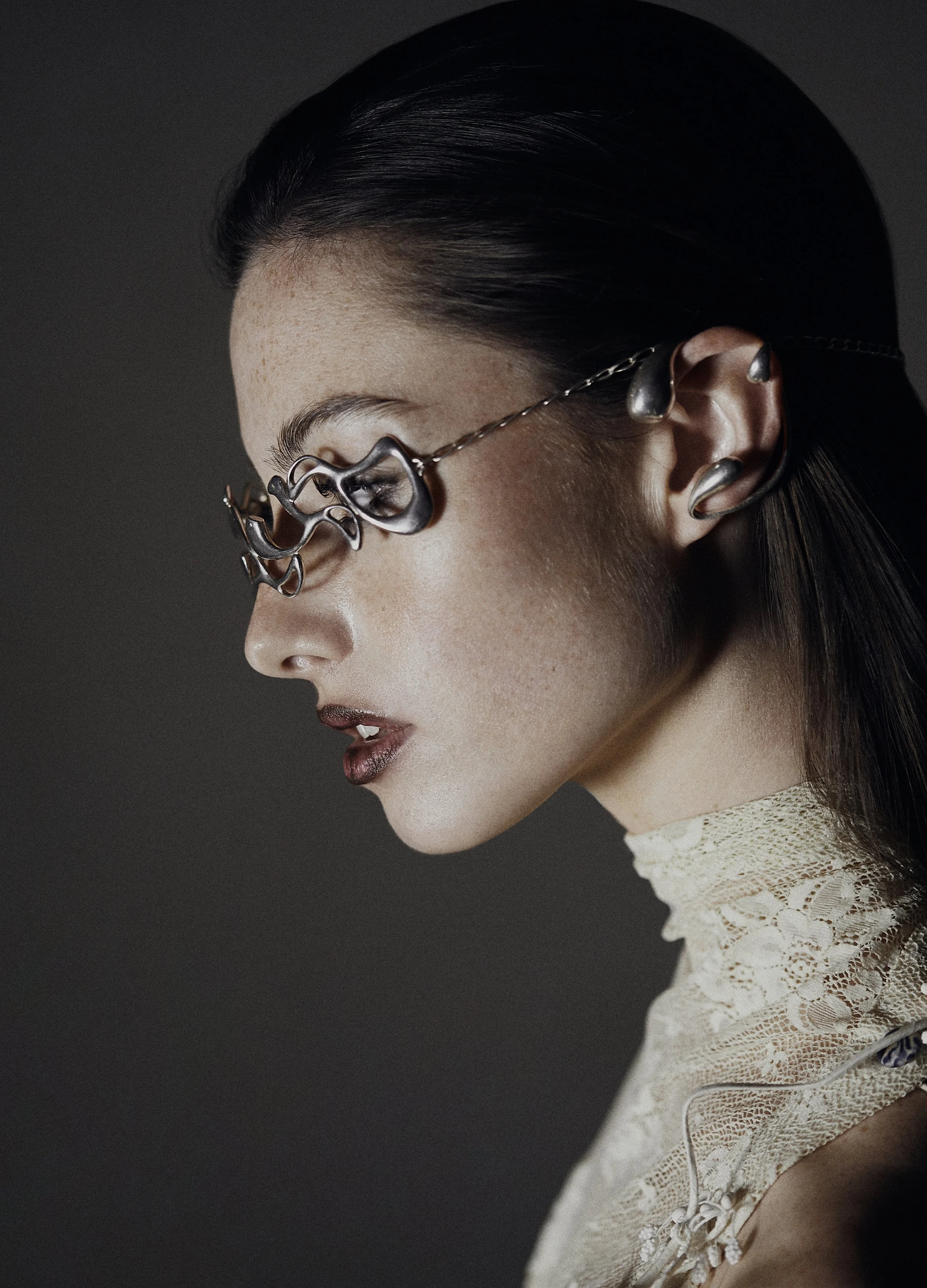 A woman with dark hair, wearing artistic metal glasses and earrings, in a side profile against a dark background.