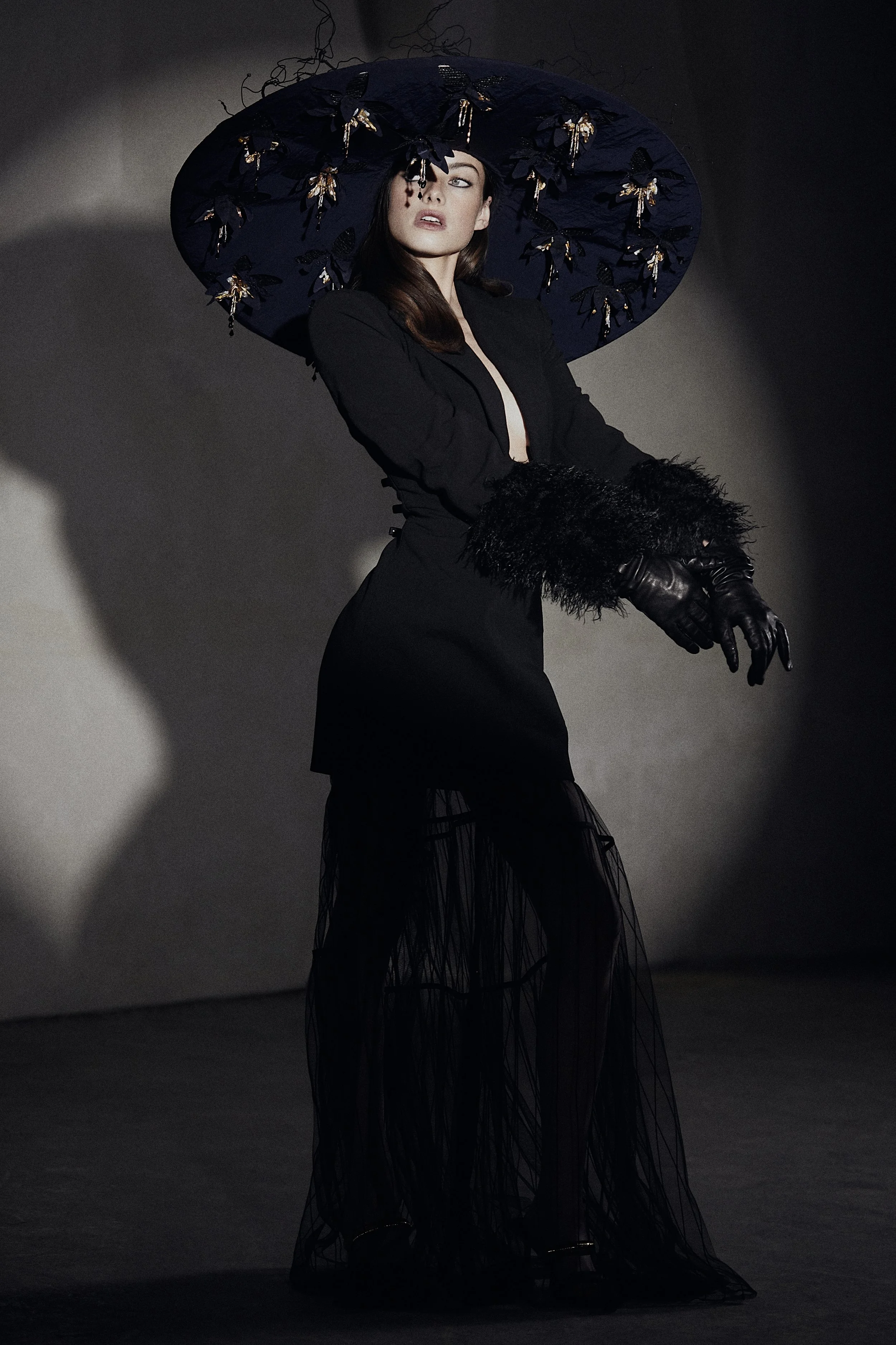 A woman dressed in black with black gloves, wearing a large navy hat decorated with black and gold butterflies, posing against a dark background.