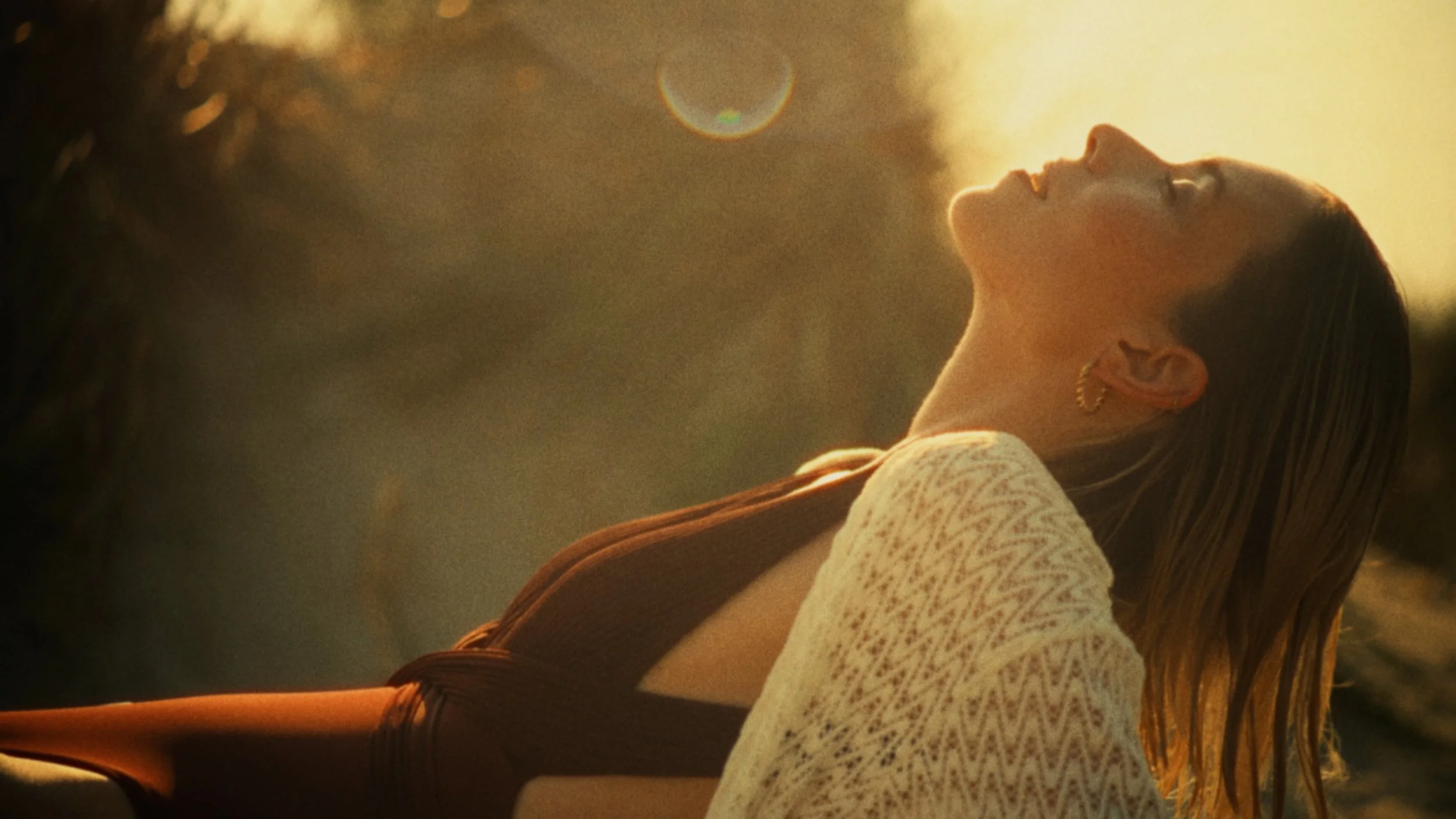A woman with wet hair and gold hoop earrings is leaning back with her eyes closed, bathed in warm sunlight, wearing a beige sweater over a brown top.