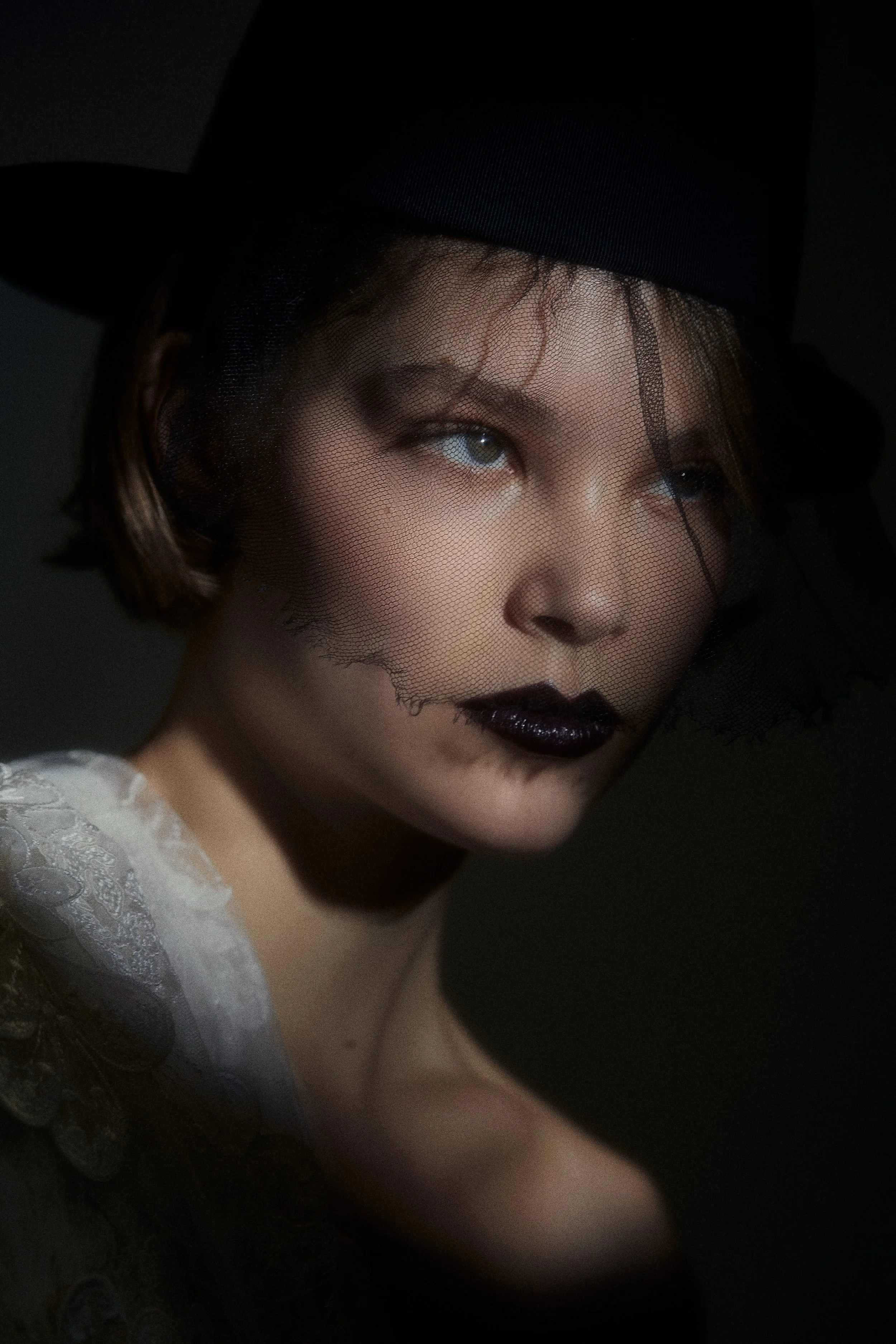 Close-up of a person with a black hat with veil, dark lipstick, and minimal makeup, wearing a white lace top, against a dark background.