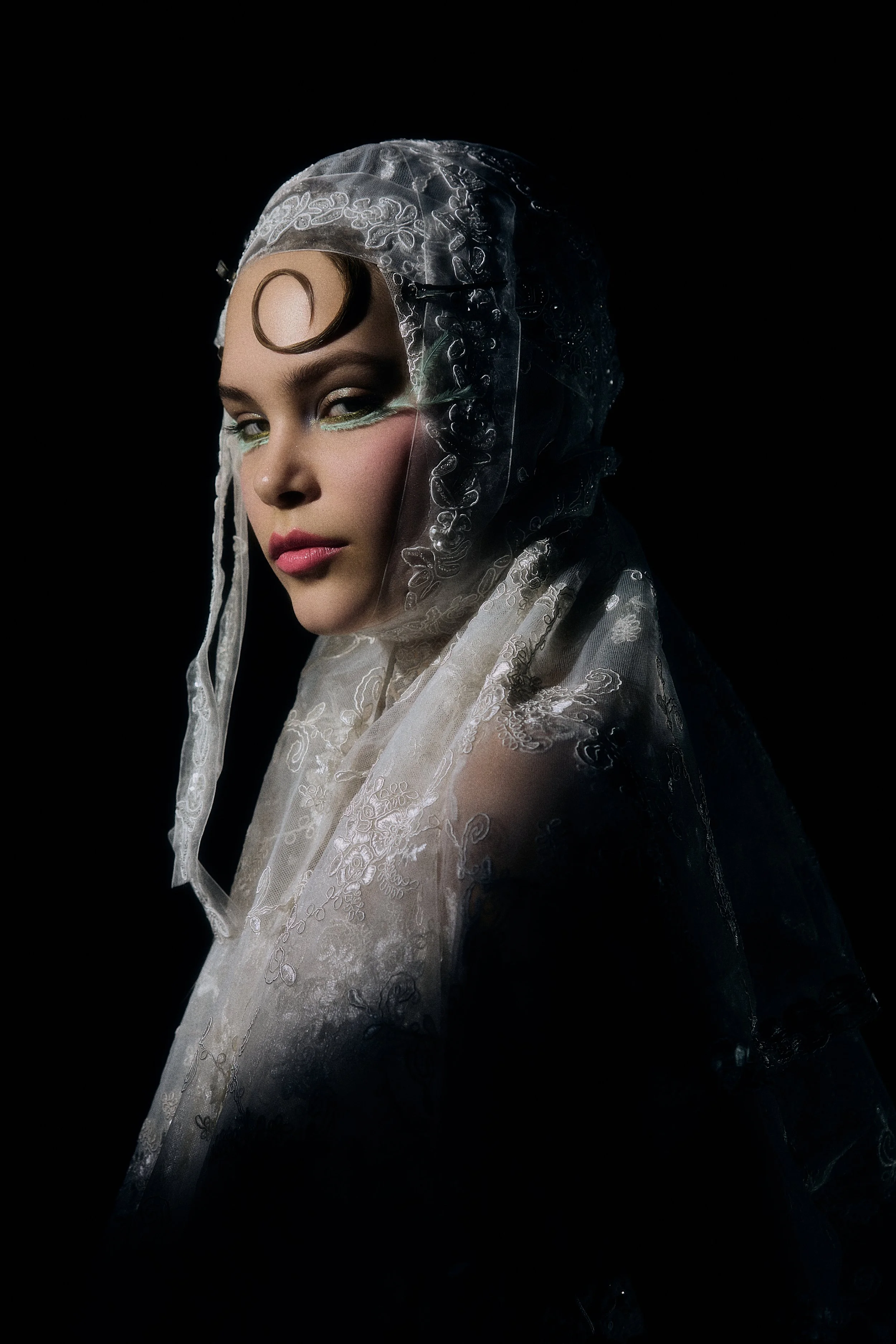 Portrait of a woman with styled hair and makeup wearing a sheer lacy head covering against a black background.