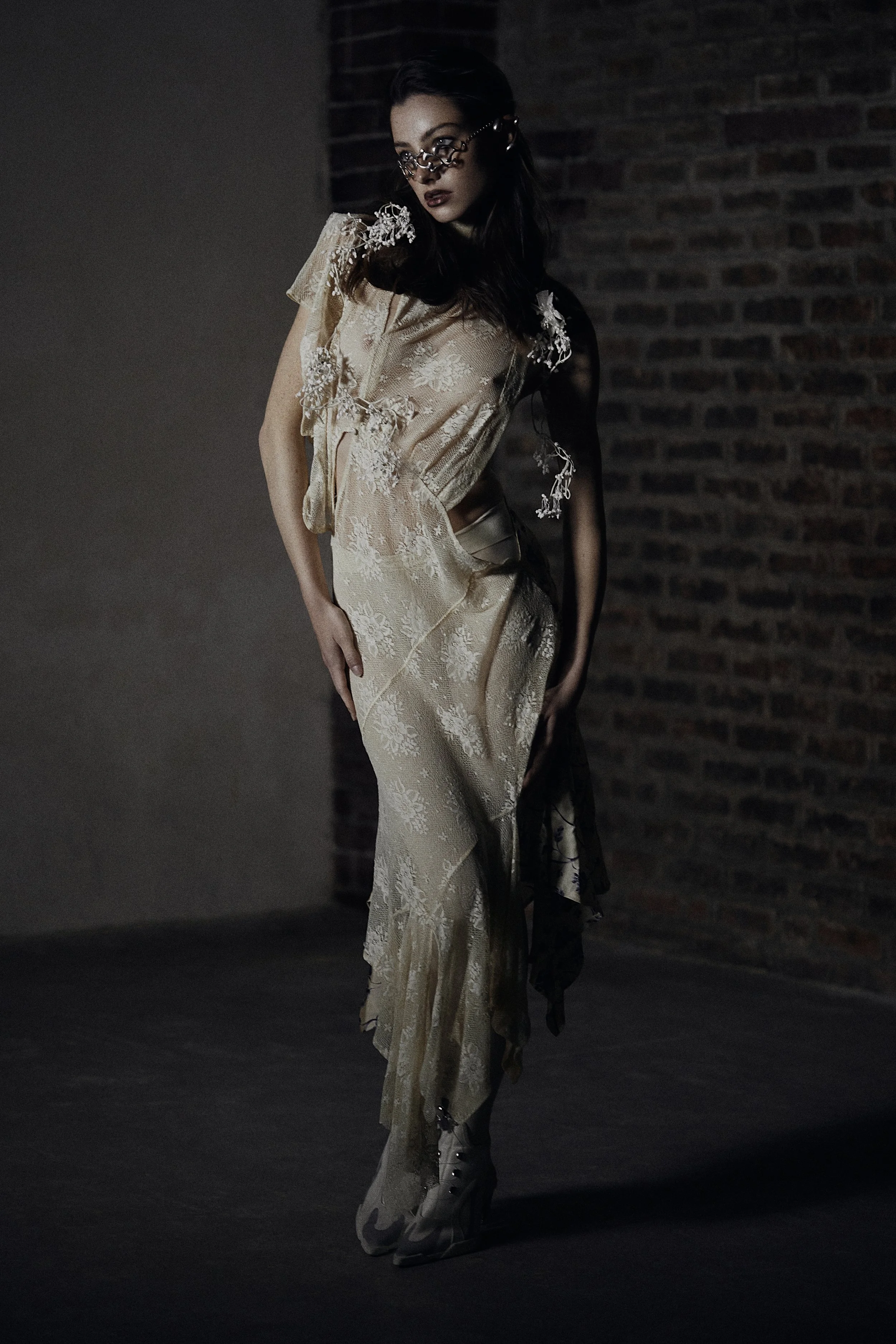 A woman wearing a beige, floral-patterned dress with lace details, paired with high fashion glasses, posed against a dark background with a brick wall.