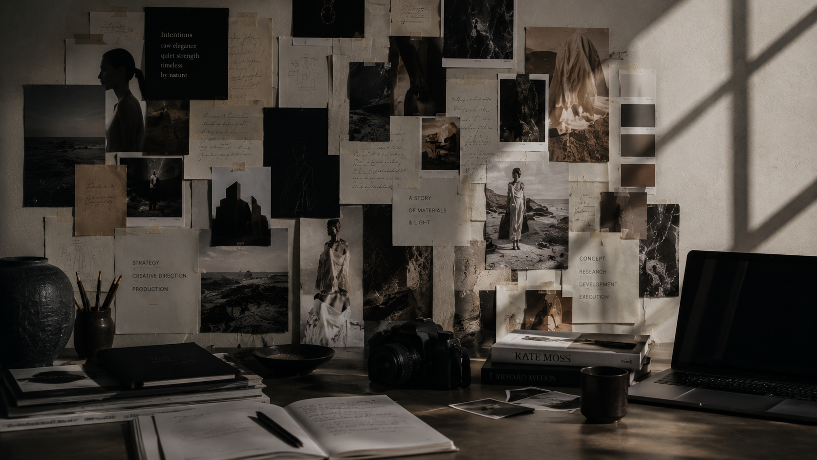 A creative workspace with a wall filled with black-and-white photographs, sketches, and notes. The desk has notebooks, a camera, a laptop, books, a cup, and drawing tools, illuminated by natural light creating shadows.