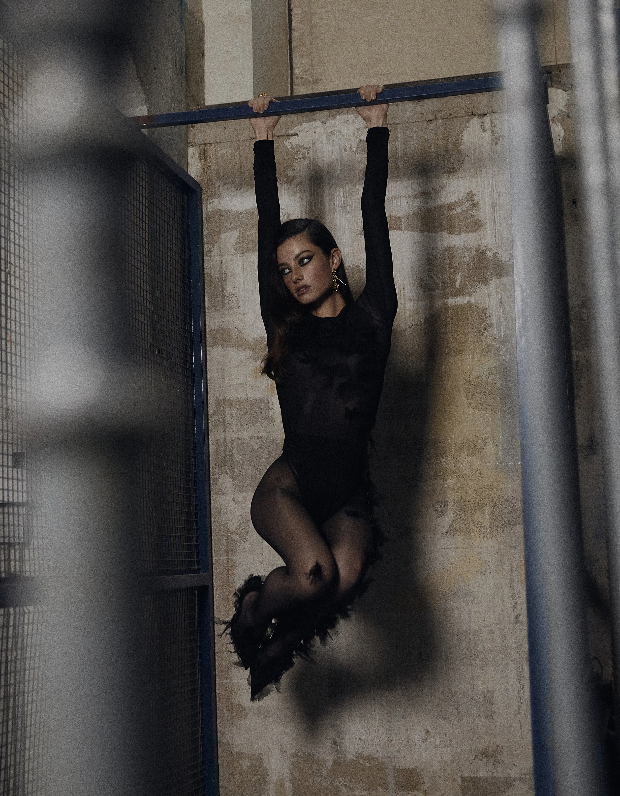 A woman dressed in black lingerie and feathered heels hanging from a pull-up bar in an industrial-style room with concrete walls.